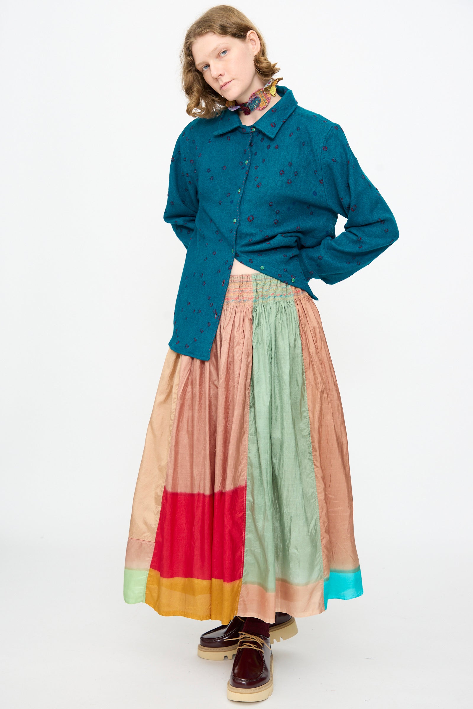 A person wearing a teal button-up shirt, the Injiri Clamp Dyed Silk Cotton Alisha Skirt in Multi featuring delicate hand embroidery, maroon platform shoes, and a floral neck scarf stands against a plain white background.