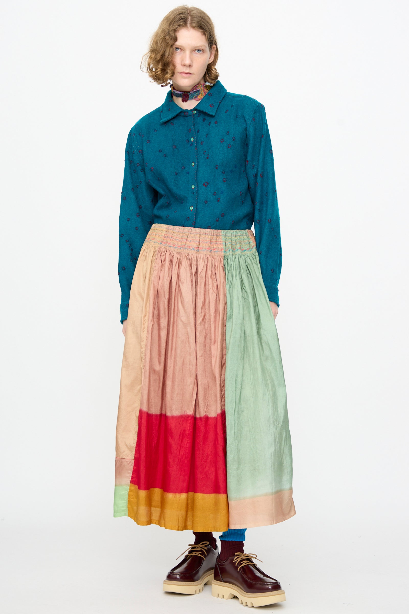 A person stands against a white background, wearing a teal button-up shirt paired with the Injiri Clamp Dyed Silk Cotton Alisha Skirt in Multi—a vibrant maxi skirt featuring red, pink, green, and yellow panels crafted from a soft cotton silk blend—and brown shoes.