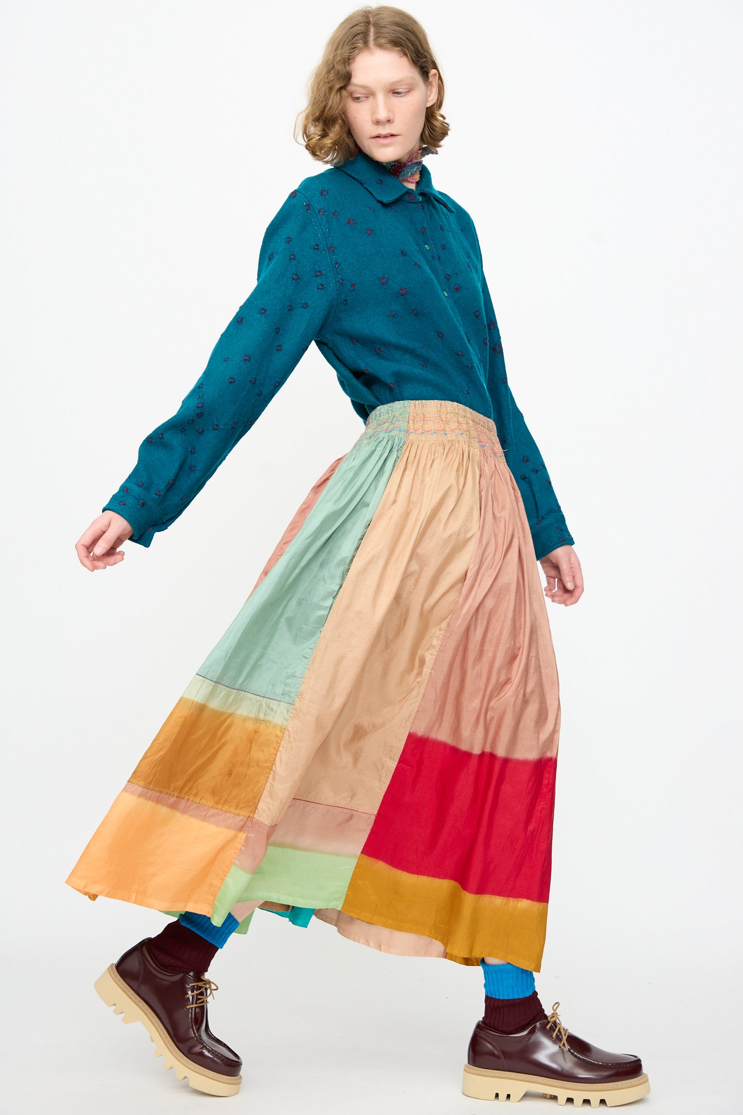 Person wearing a teal shirt with small patterns, the Injiri Clamp Dyed Silk Cotton Alisha Skirt in Multi—a maxi skirt crafted from a luxurious cotton-silk blend featuring multicolored patchwork—paired with teal socks and brown platform shoes, posing against a plain white background.