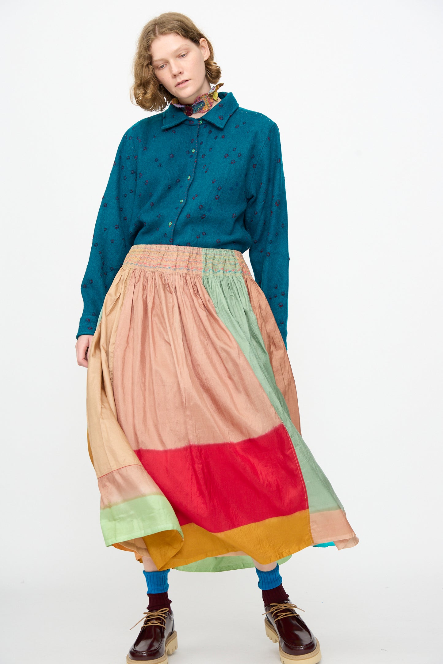 A person wearing a teal button-up shirt with floral embroidery, paired with the Injiri Clamp Dyed Silk Cotton Alisha Skirt in Multi and brown shoes, stands against a plain white background.