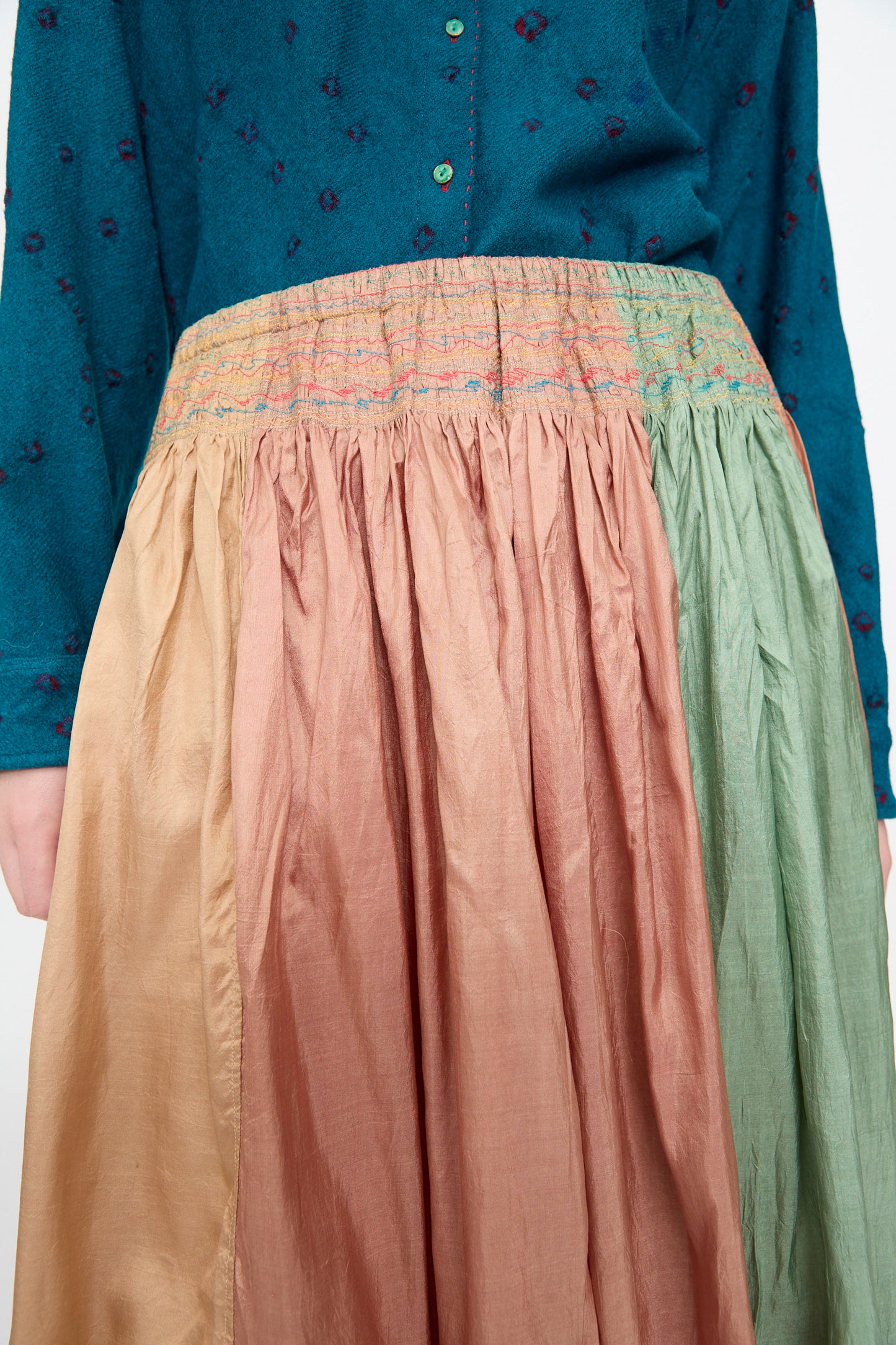 Person wearing a teal long-sleeve shirt and the Injiri Clamp Dyed Silk Cotton Alisha Skirt in Multi—a gathered, color-blocked maxi skirt featuring gold, pink, and green panels, crafted from a luxurious cotton silk blend.