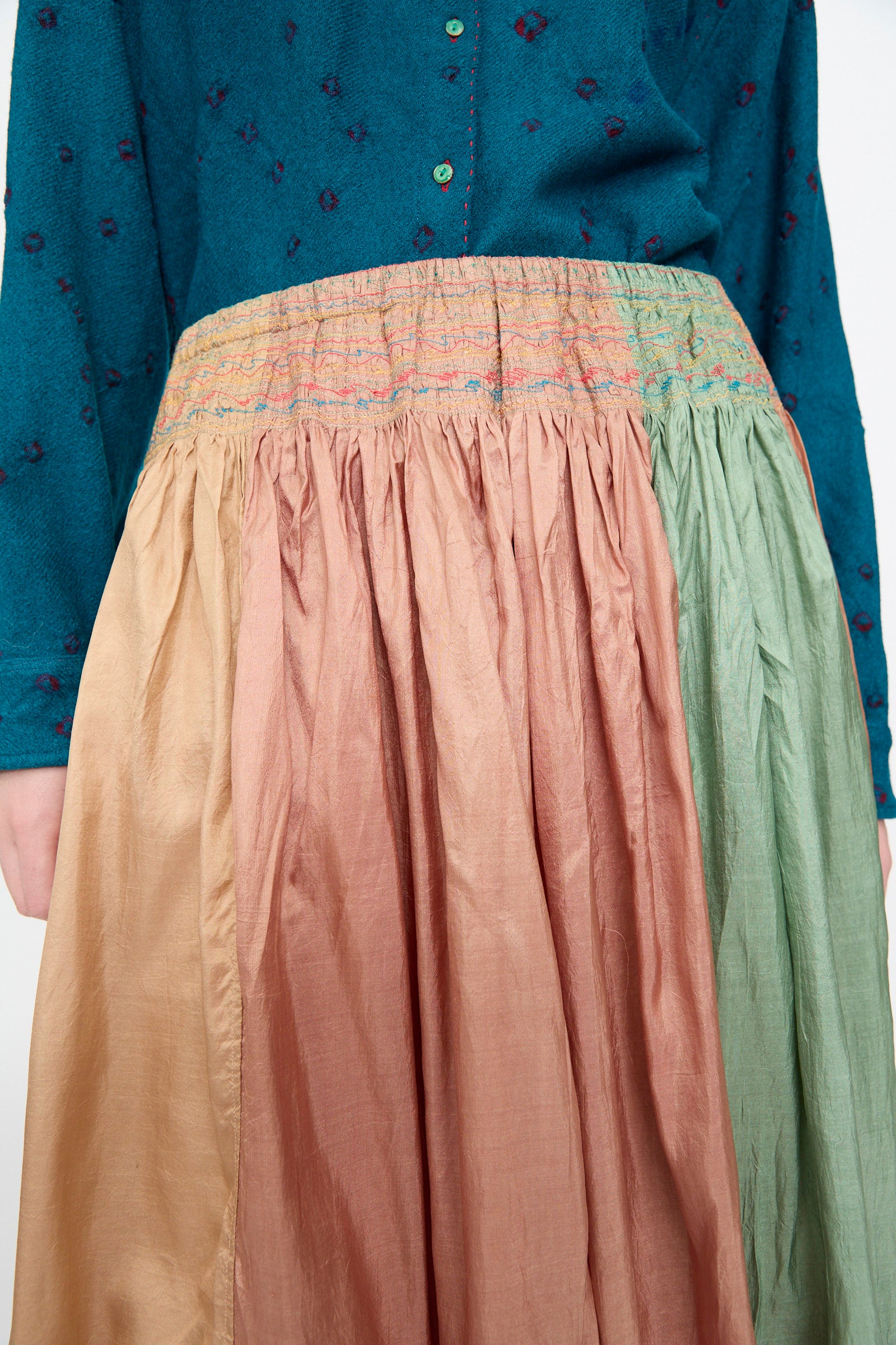 Person wearing a teal long-sleeve shirt and the Injiri Clamp Dyed Silk Cotton Alisha Skirt in Multi—a gathered, color-blocked maxi skirt featuring gold, pink, and green panels, crafted from a luxurious cotton silk blend.
