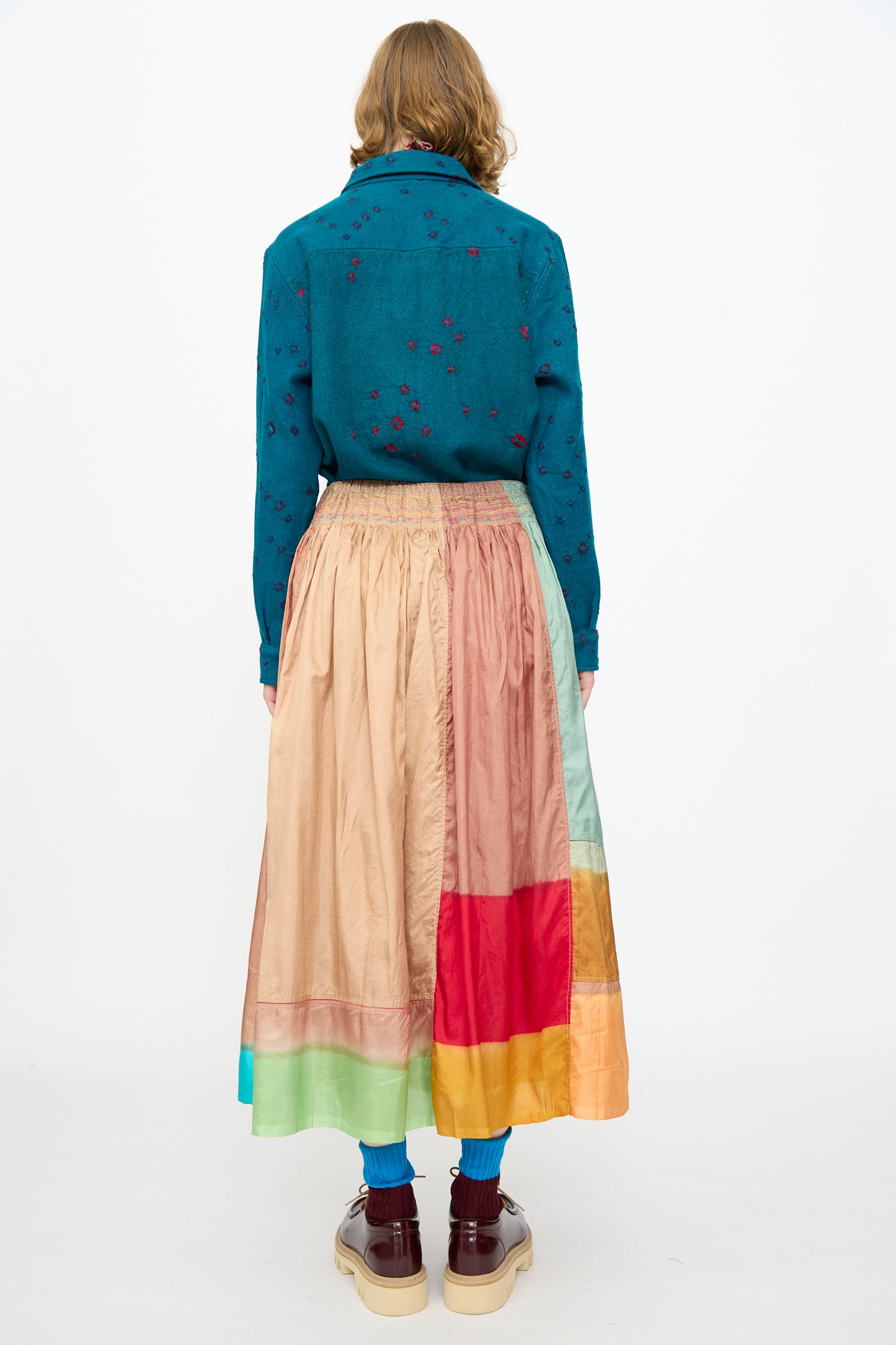 A person stands with their back to the camera against a plain white background, wearing a teal shirt with small patterns crafted from a cotton silk blend, paired with the Injiri Clamp Dyed Silk Cotton Alisha Skirt in Multi, blue socks, and brown platform shoes.