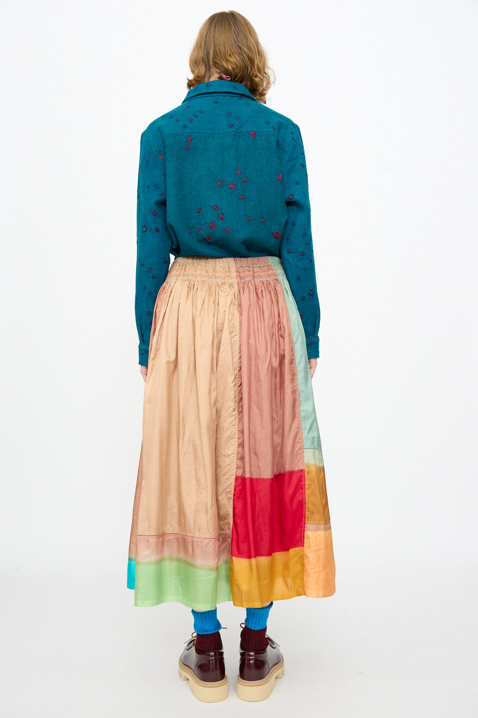 A person stands with their back to the camera against a plain white background, wearing a teal shirt with small patterns crafted from a cotton silk blend, paired with the Injiri Clamp Dyed Silk Cotton Alisha Skirt in Multi, blue socks, and brown platform shoes.