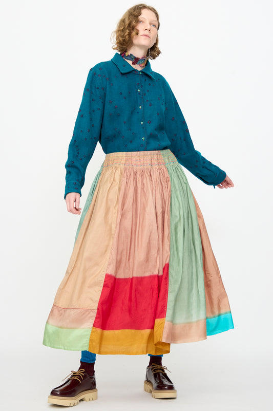 A person wearing a teal button-up shirt with the Injiri Clamp Dyed Silk Cotton Alisha Skirt in Multi—a long, color-blocked maxi skirt crafted from a silk-cotton blend and adorned with delicate hand embroidery—and brown platform shoes, standing against a plain white background.