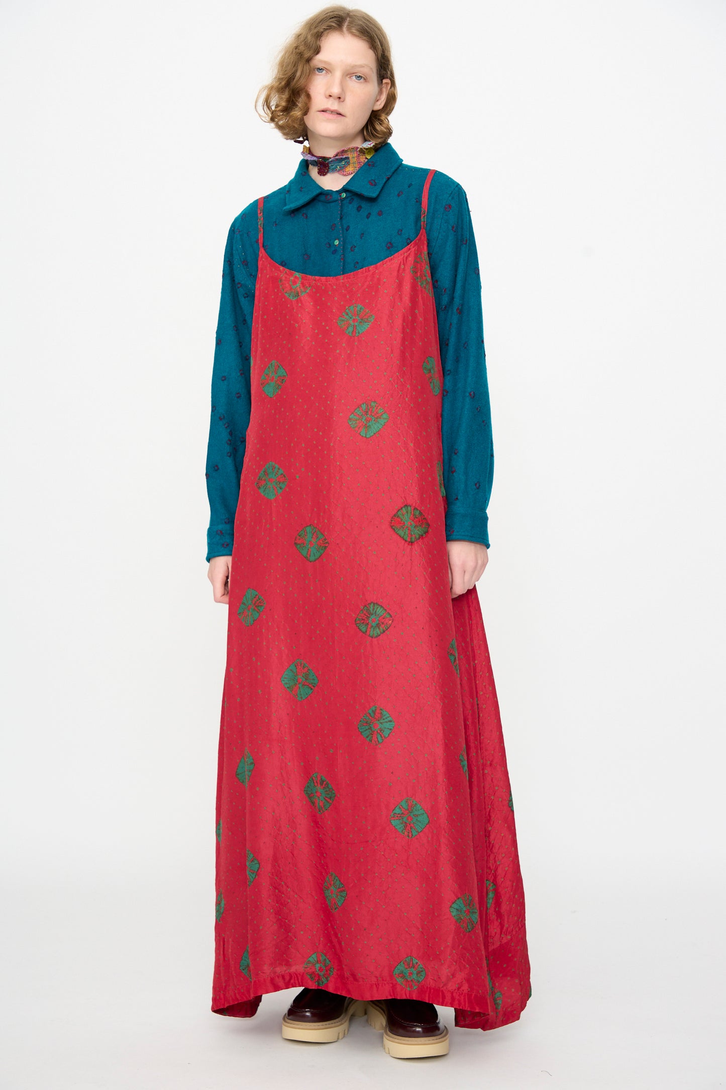 A person wears the Injiri Cotton Bandhani Silk Slip in Red—a long red maxi dress with green patterns—layered over a blue button-up shirt, standing against a plain white background.