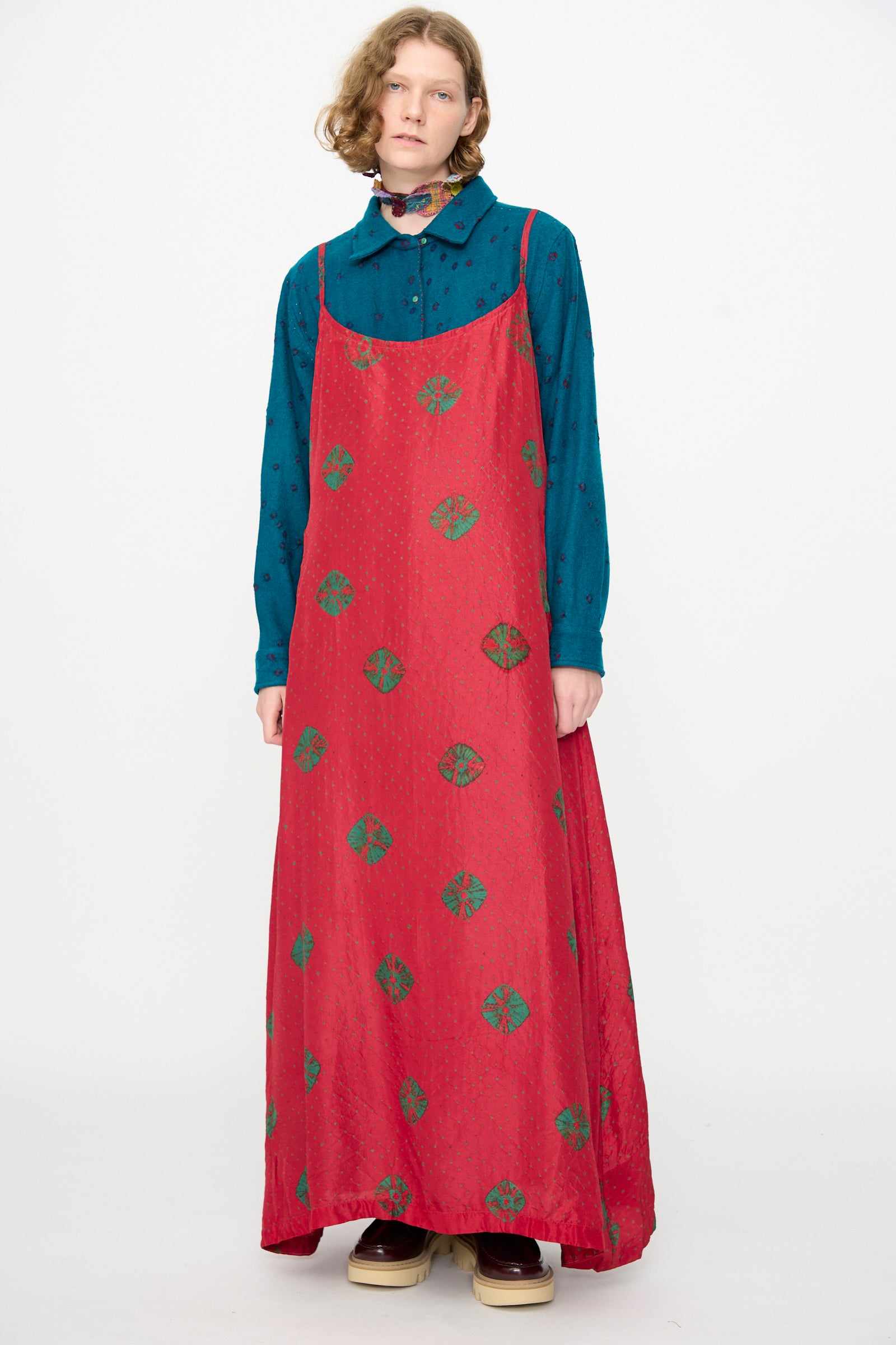 A person wears the Injiri Cotton Bandhani Silk Slip in Red—a long red maxi dress with green patterns—layered over a blue button-up shirt, standing against a plain white background.