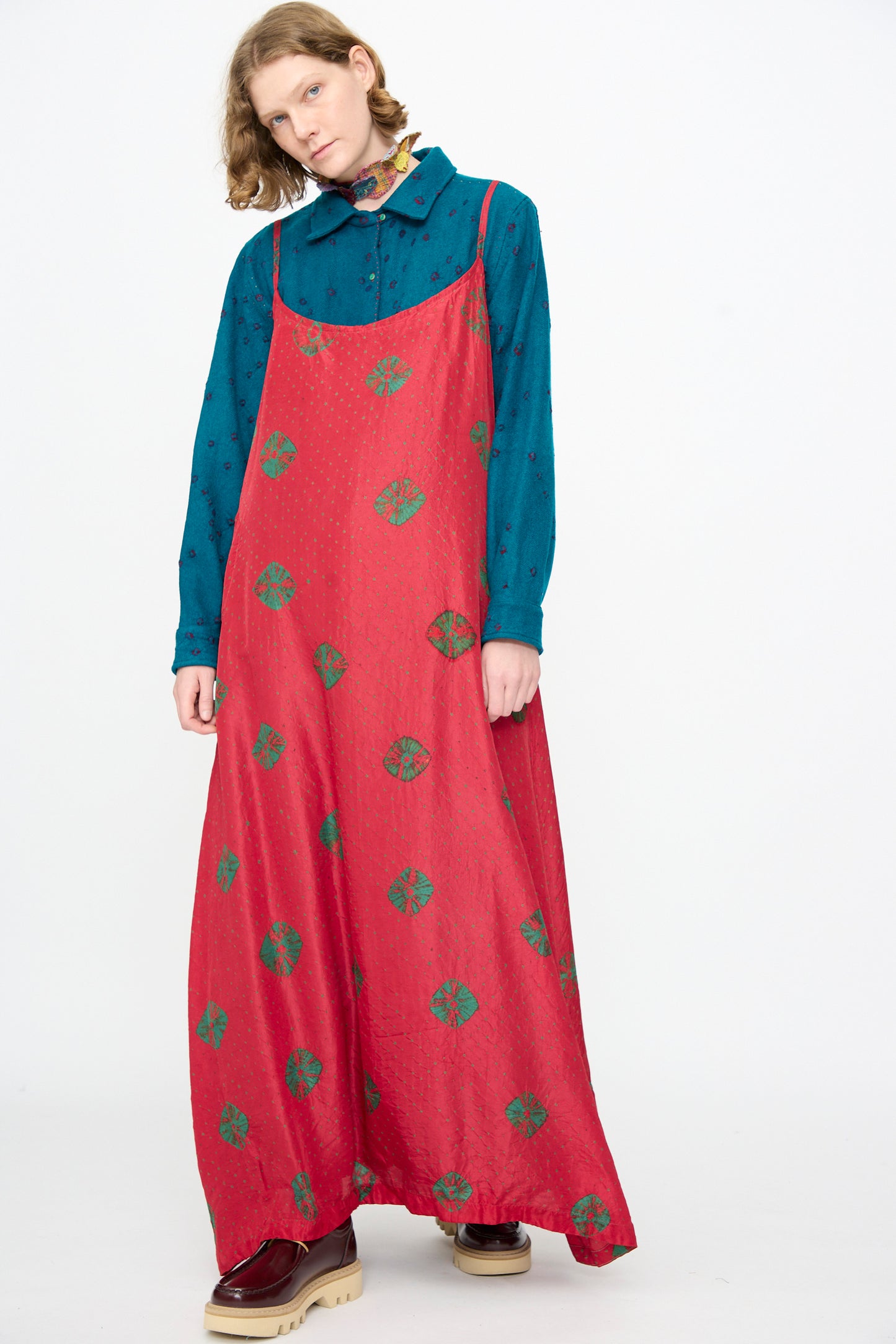 A person is wearing the Injiri Cotton Bandhani Silk Slip in Red—a long red maxi slip dress with green circular patterns—layered over a teal collared shirt and paired with red platform shoes, standing against a plain white background.