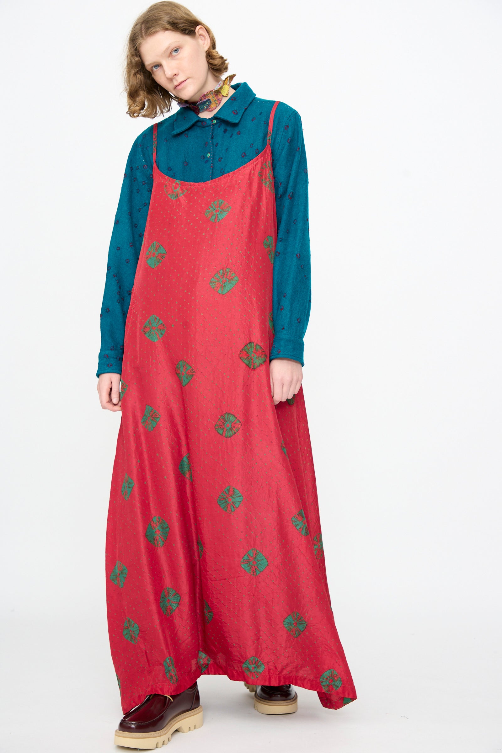 A person is wearing the Injiri Cotton Bandhani Silk Slip in Red—a long red maxi slip dress with green circular patterns—layered over a teal collared shirt and paired with red platform shoes, standing against a plain white background.