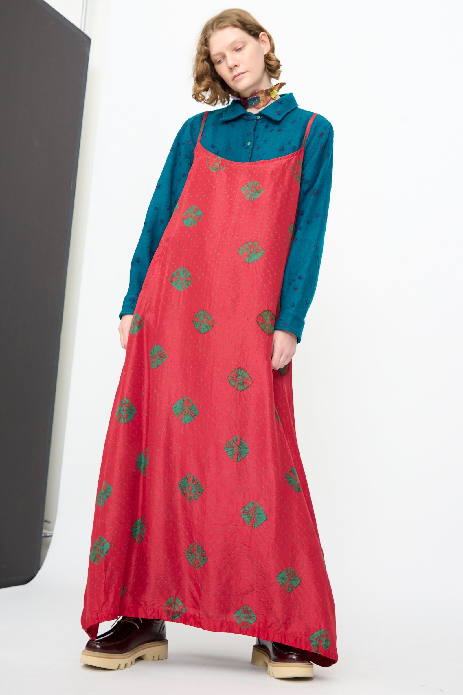 A person is wearing the “Cotton Bandhani Silk Slip in Red” by Injiri, featuring green Bandhani tie-dye patterns, layered over a blue collared shirt and paired with beige platform shoes, standing against a plain background.