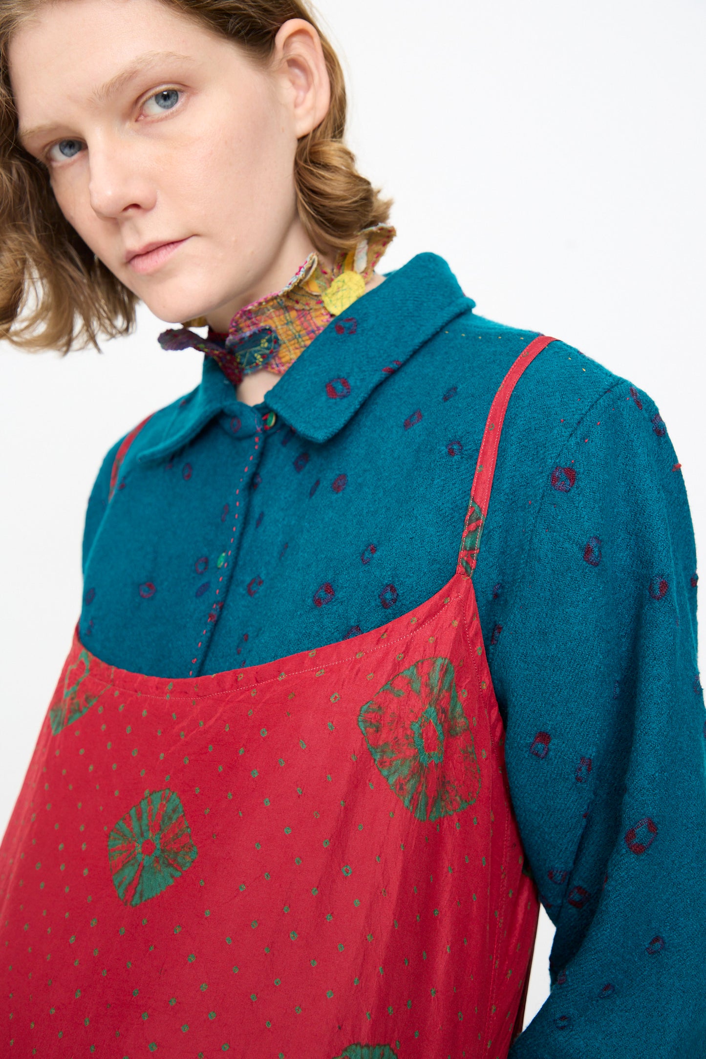 A person with short light brown hair wears a teal collared shirt layered under the Injiri Cotton Bandhani Silk Slip in Red, styled with a colorful neck scarf, standing against a plain background.