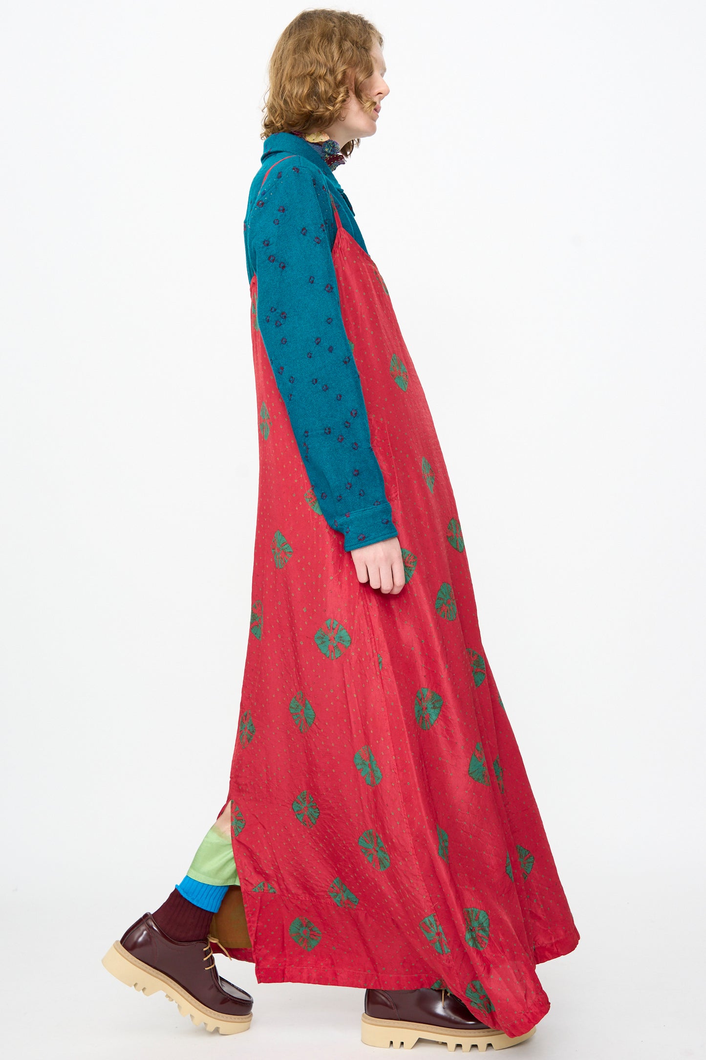 A person walks against a white background wearing a teal long-sleeve shirt layered under the Injiri Cotton Bandhani Silk Slip in Red, paired with maroon shoes and mismatched socks.