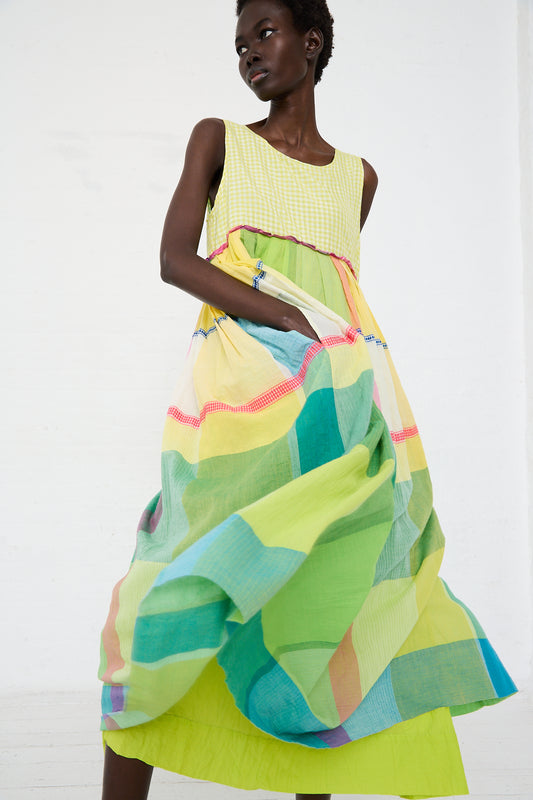 A Black woman models a colorful, flowing empire waist Cotton Check Gingham Dress in Green, Yellow and Pink with diagonal stripes in a bright, white studio space by Injiri.