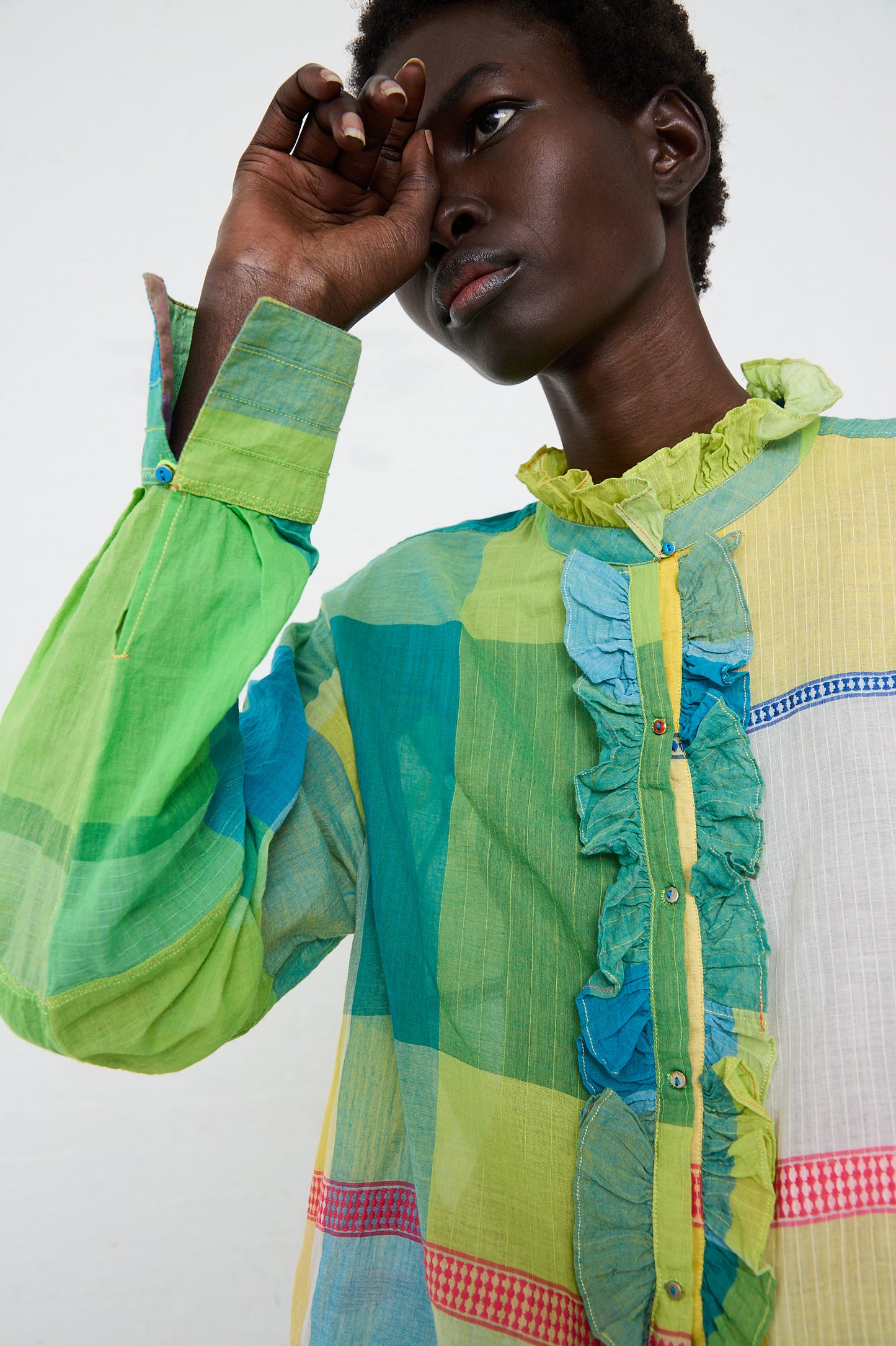 A woman in an Injiri cotton shirt in green, yellow and pink check ponderingly touches front of her head, against a white background.