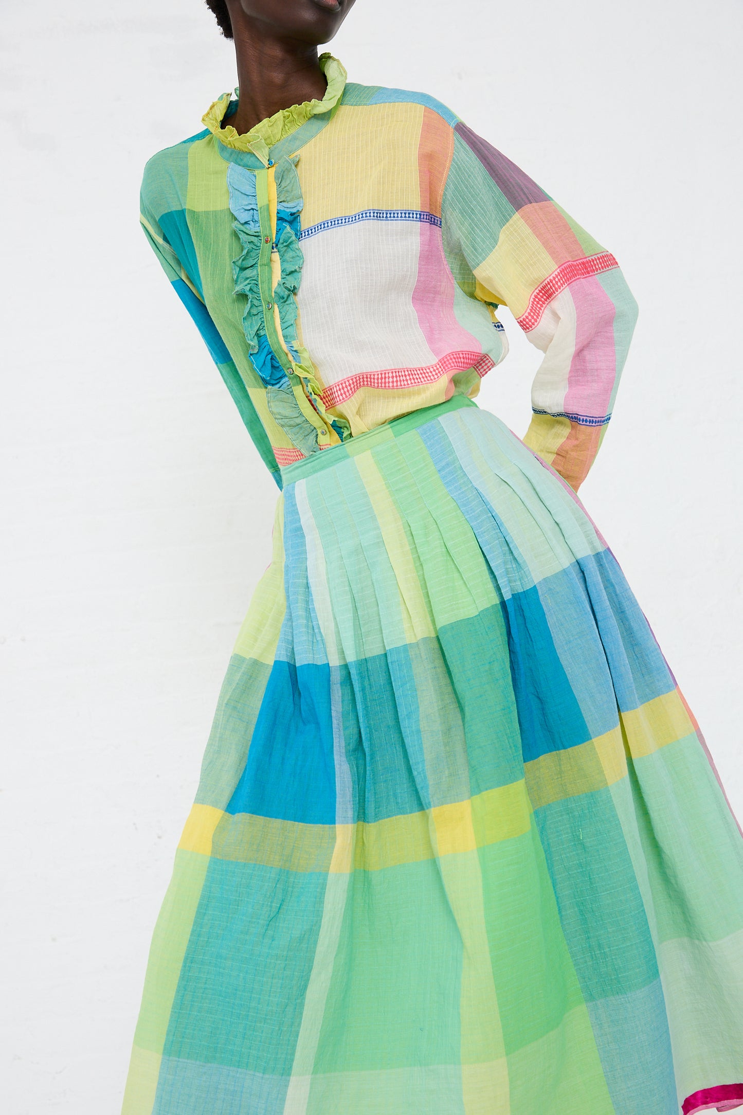 A person models an Injiri Cotton Shirt in Green, Yellow and Pink Check with a ruffled neckline and pleated skirt, standing against a white background.