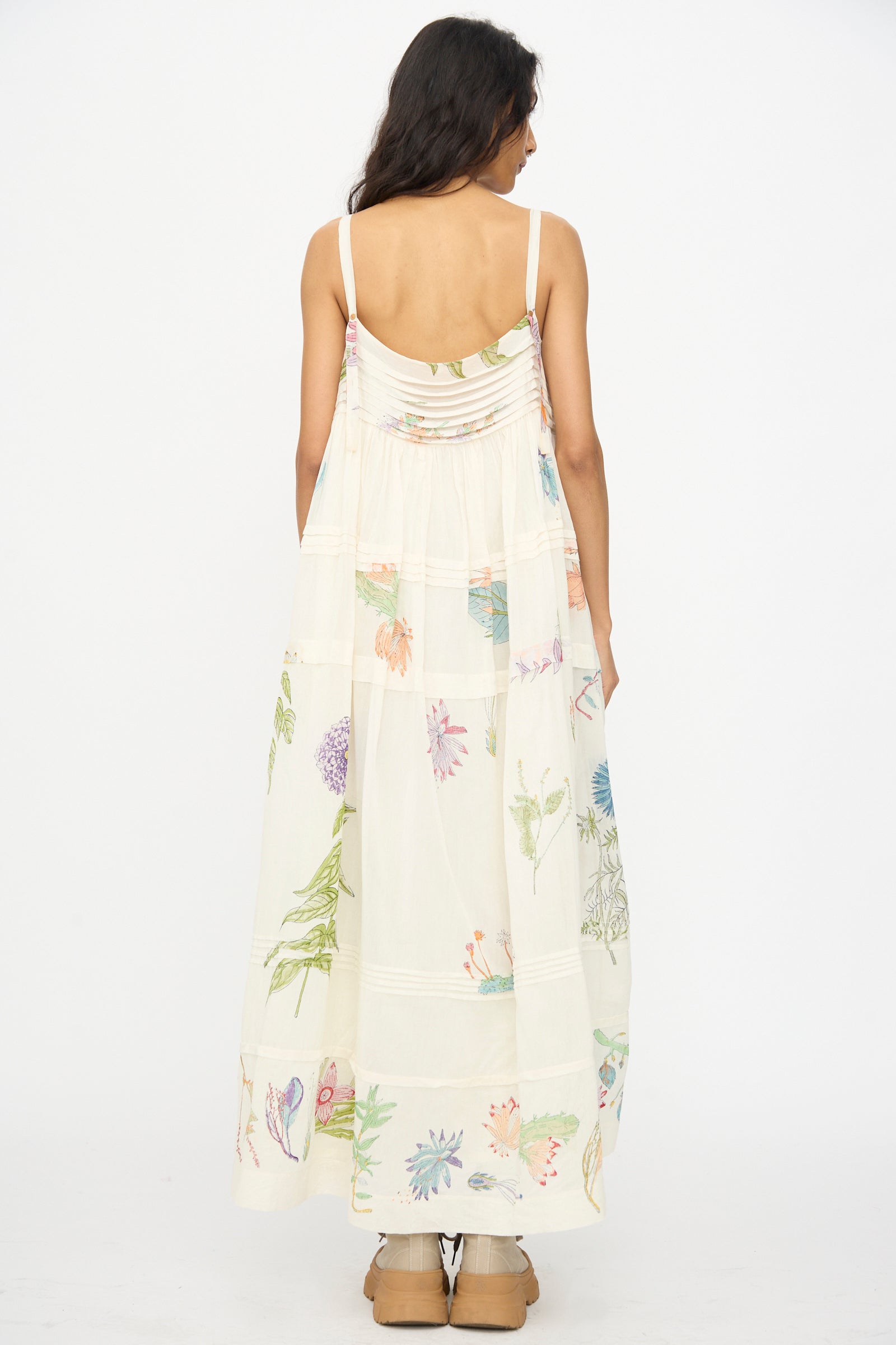 A woman with long dark hair stands with her back to the camera against a white background, wearing Injiri's Cotton Silk Blockprint Hazel Slip in White Floral and tan shoes.