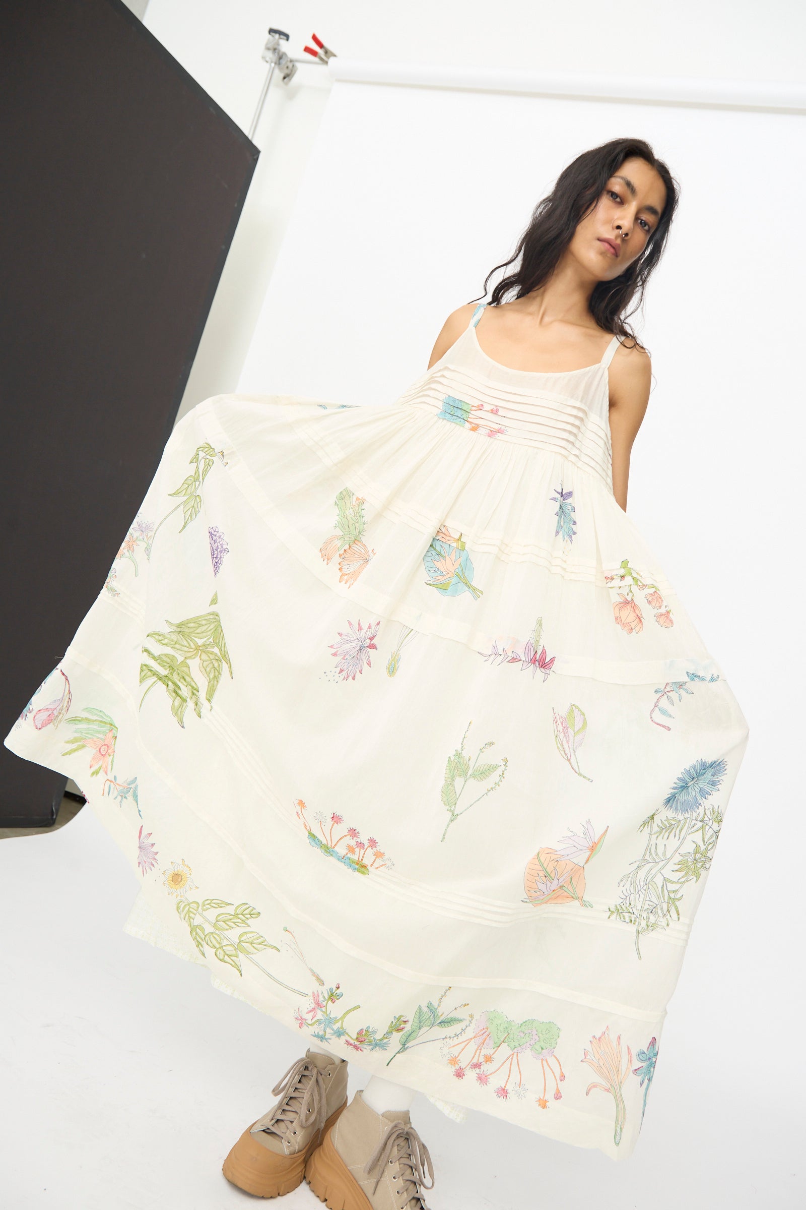 A woman wears the Injiri Cotton Silk Blockprint Hazel Slip in White Floral, holding out the skirt while posing in a studio with a white background and tan boots.
