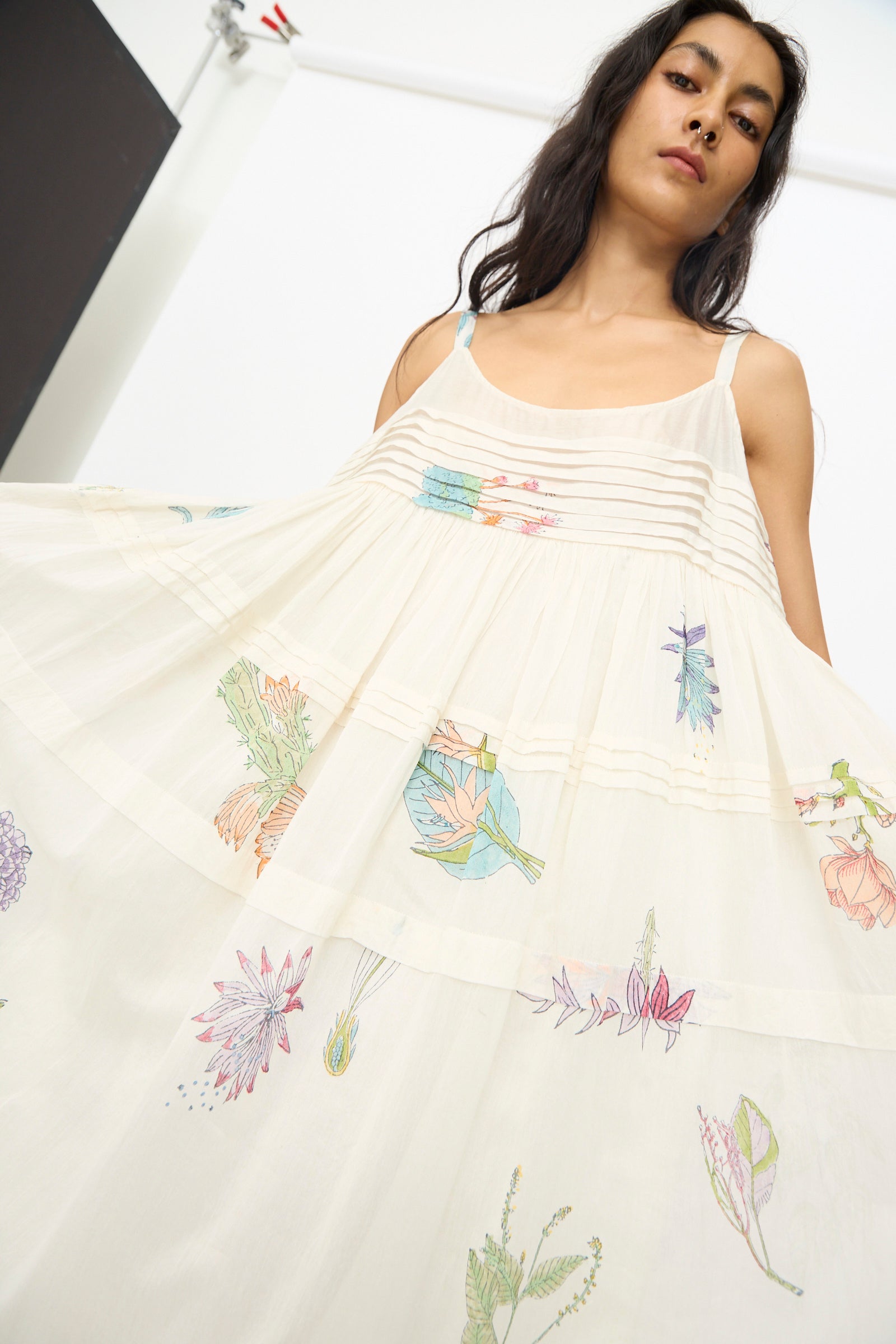 A woman gracefully holds out the skirt of the Injiri Cotton Silk Blockprint Hazel Slip in White Floral, featuring pleated details and colorful botanical embroidery.