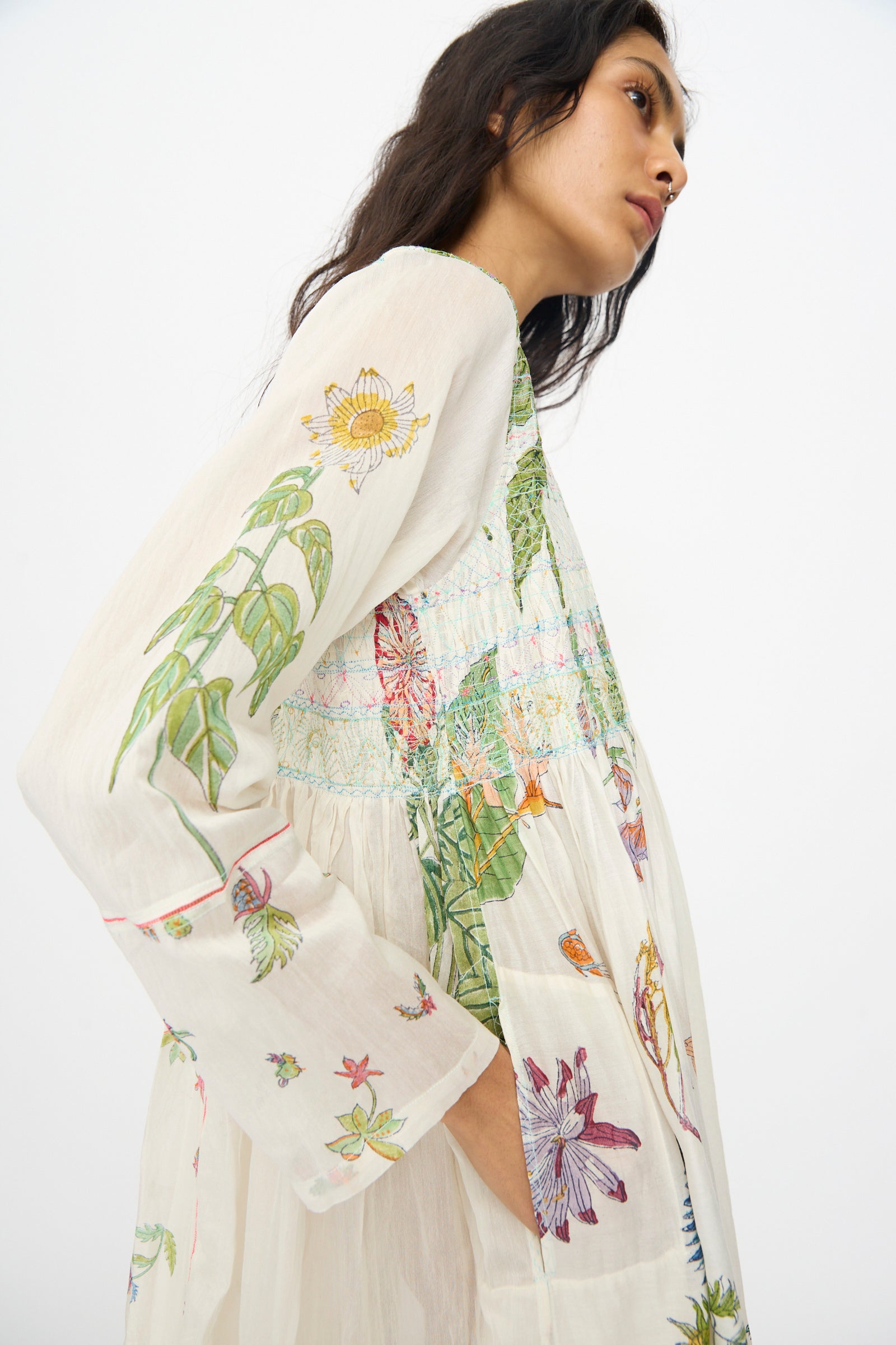 A woman wears the Injiri Cotton Silk Blockprint Nazma Dress in White, featuring colorful floral print and long sleeves, as she stands against a plain white background, gazing thoughtfully to the side.