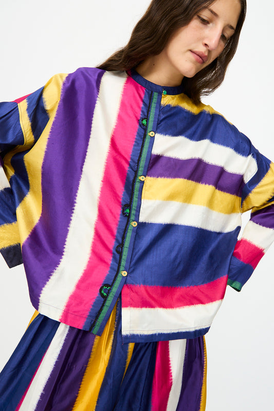 A woman wears the Injiri Ikat Silk Shirt in Multi Stripe, featuring a loose, boxy fit and bold stripes in purple, yellow, white, pink, and blue.