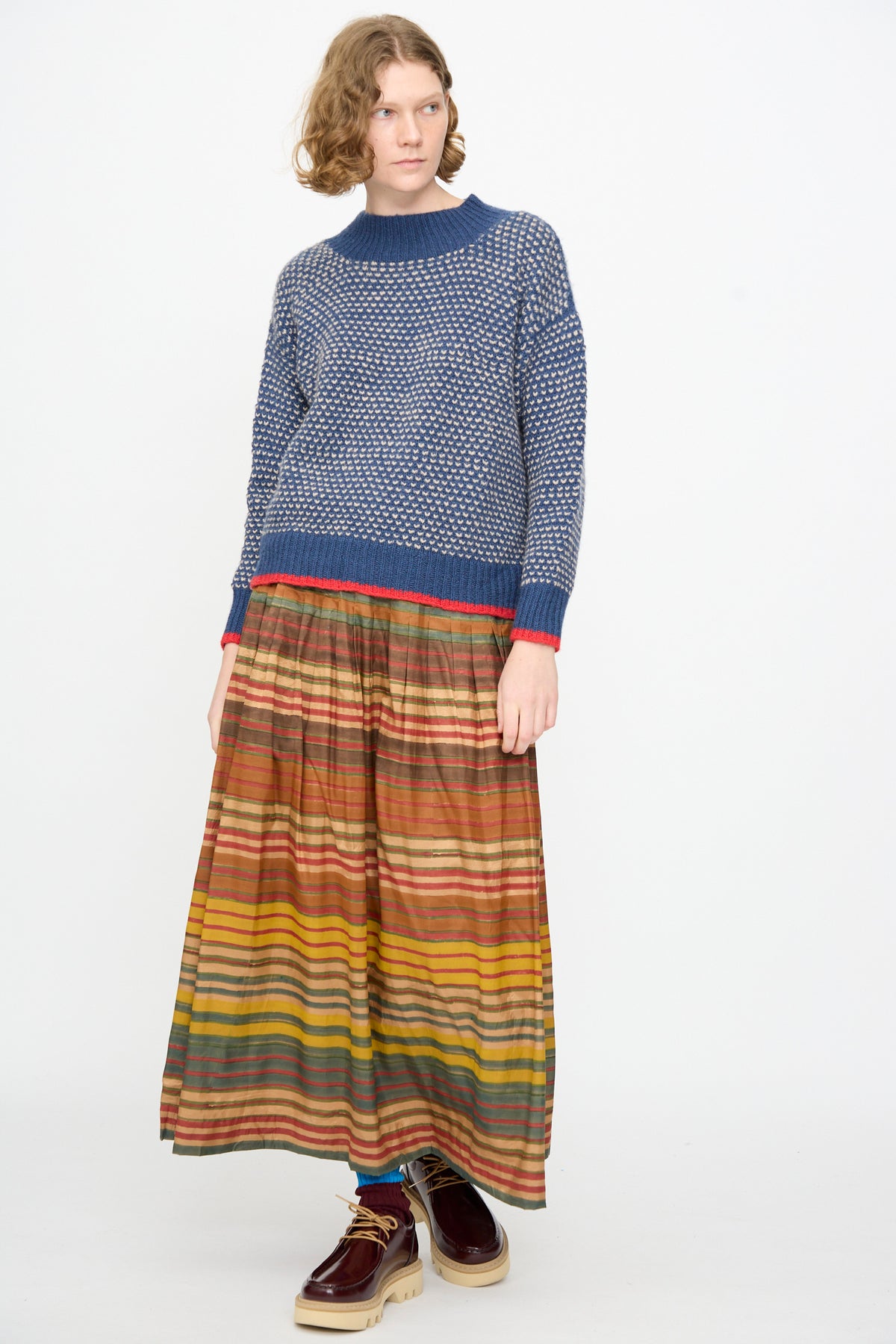 A model wears the Injiri Knitted Wool Sweater in Blue, paired with a long multicolored striped skirt and dark platform shoes, posing against a plain white background.