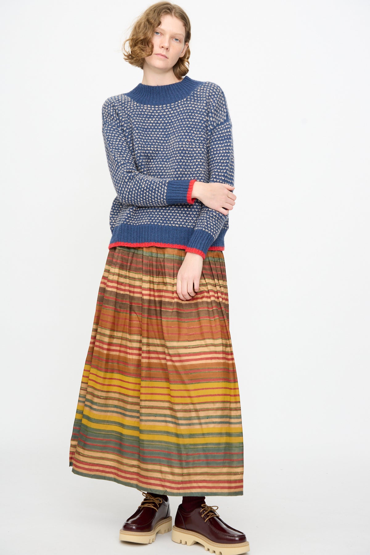 A person stands against a white background, wearing the Knitted Wool Sweater in Blue by Injiri, paired with a long multicolored striped skirt and brown platform shoes.