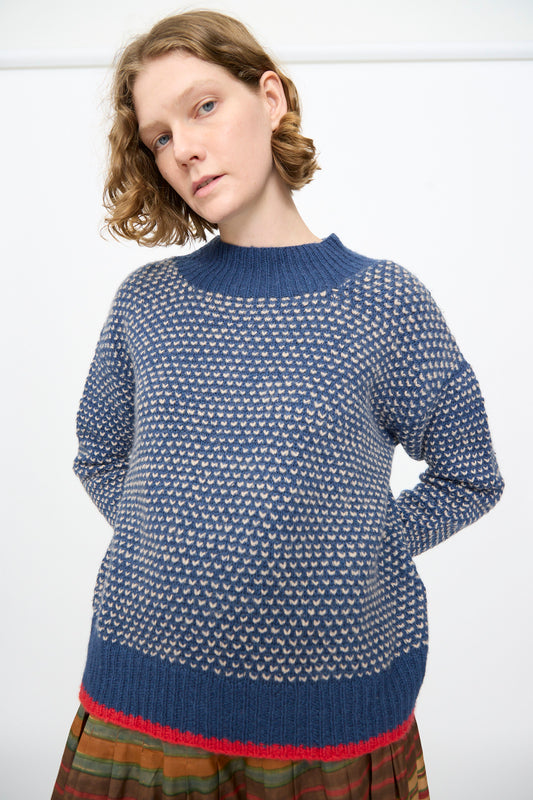 A person with short wavy hair is wearing the Knitted Wool Sweater in Blue by Injiri, paired with a multicolored skirt, standing against a plain white background.