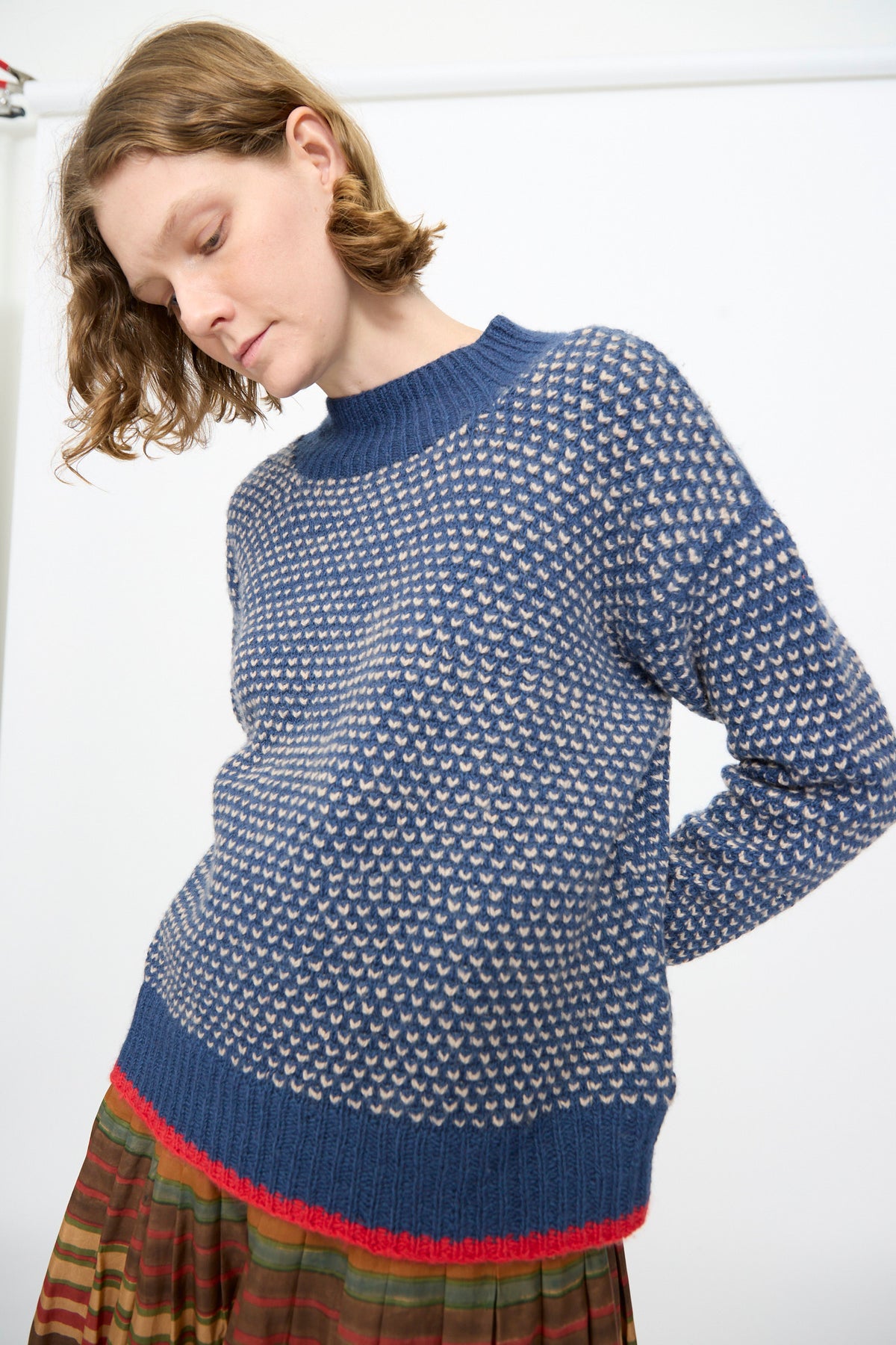 A model wearing the Injiri Knitted Wool Sweater in Blue—featuring long sleeves, a blue and white pattern, and a red stripe at the hem—paired with a multicolored striped skirt, stands against a plain white background.