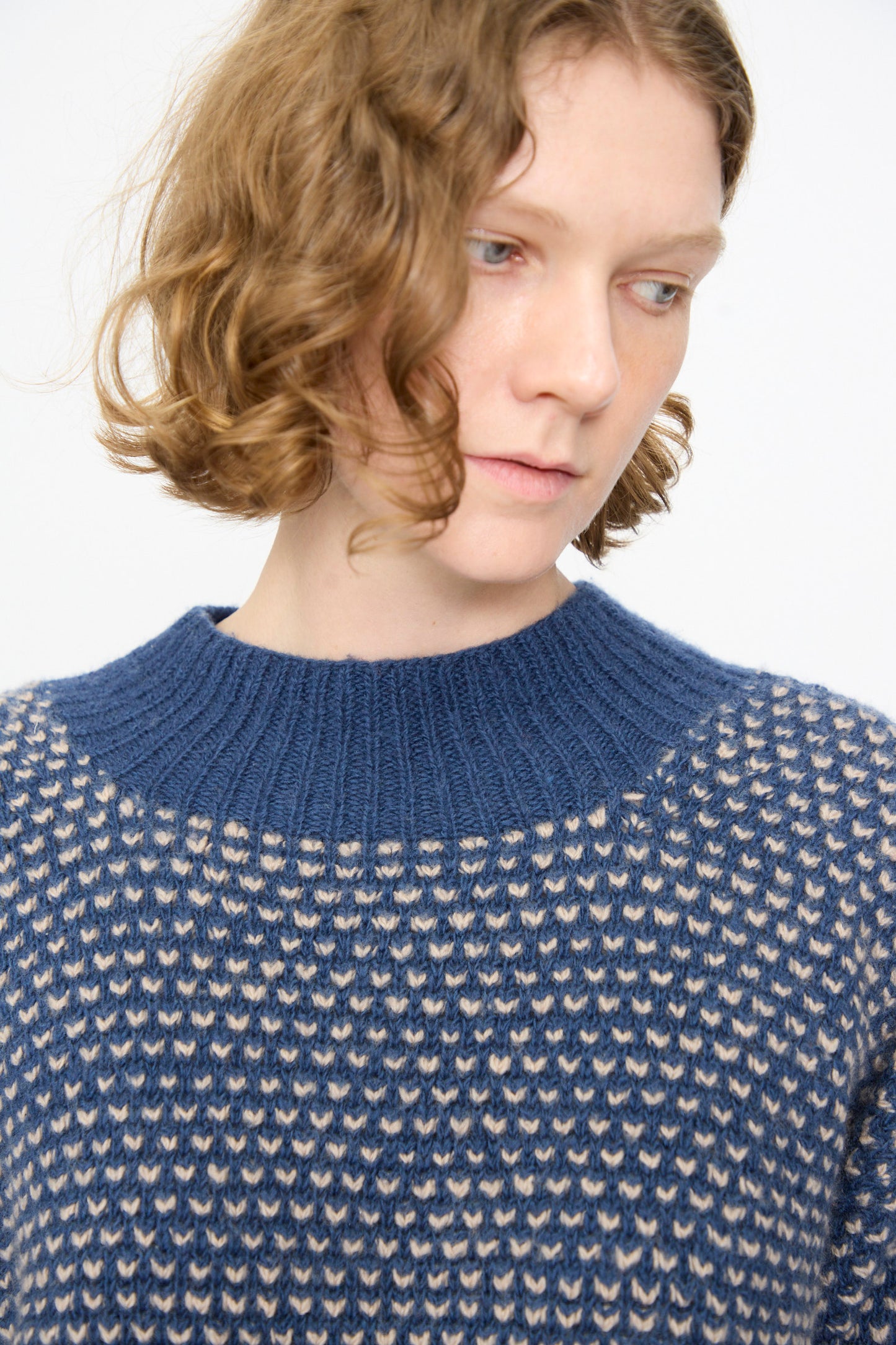 A person with short, curly light brown hair is wearing the Injiri Knitted Wool Sweater in Blue, featuring beige patterned knit details; they are looking slightly downward and to the side against a plain white background.