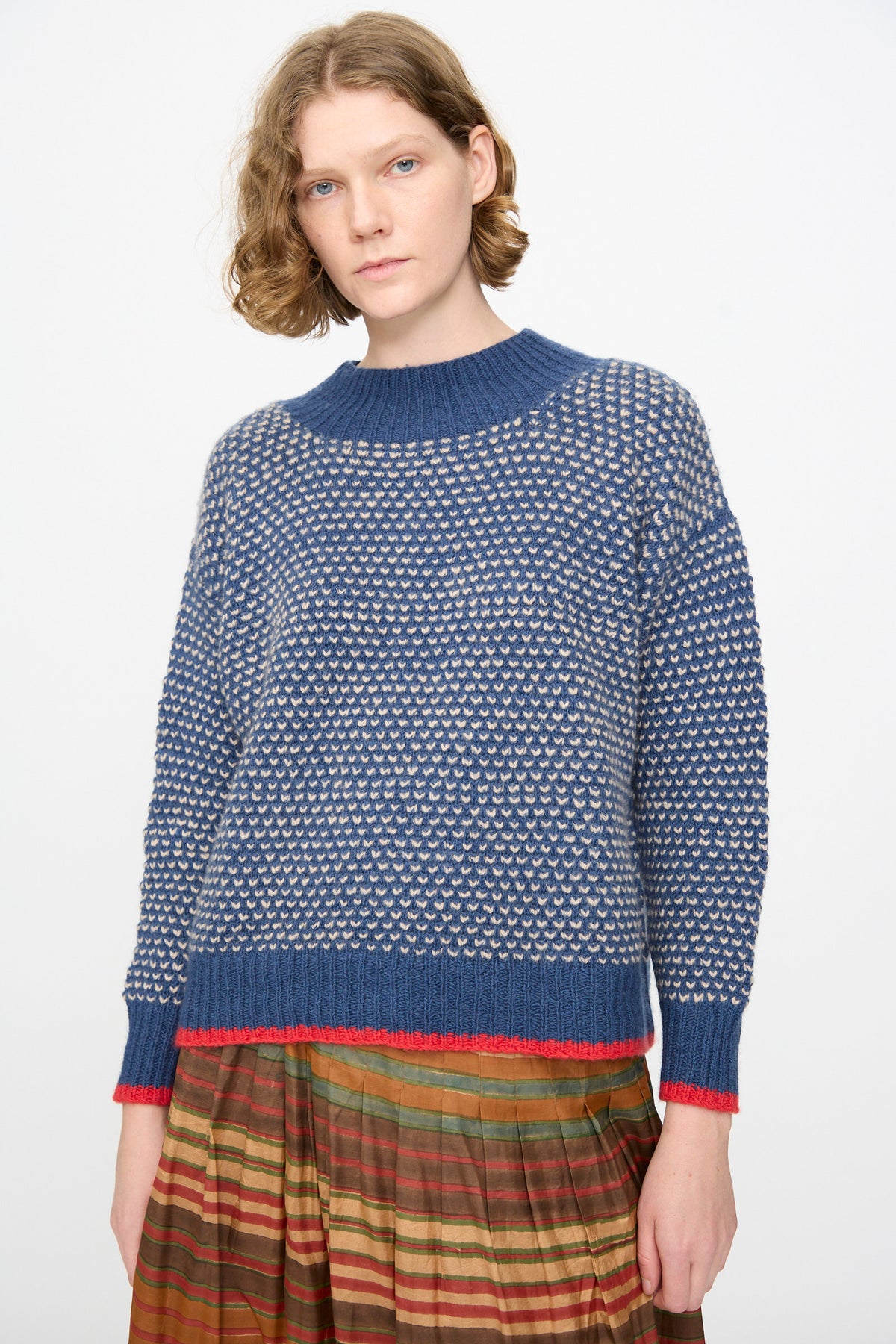 A person with short wavy hair wears the Injiri Knitted Wool Sweater in Blue, paired with a multicolored pleated skirt, standing against a plain light background.