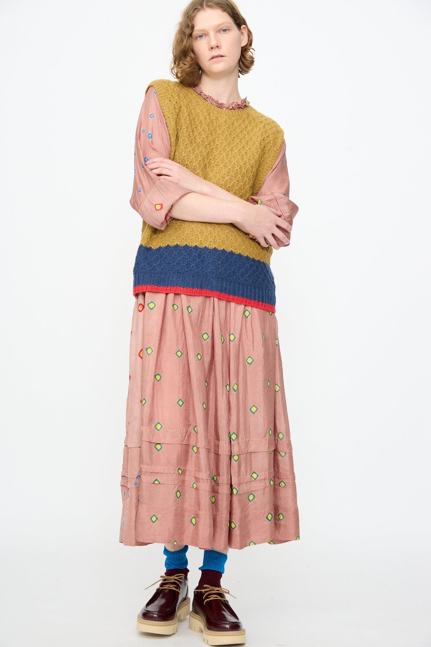 A person wears the Injiri Knitted Wool Sweater in Ochre and Blue—a boxy, textured sleeveless knit—layered over a pink embroidered dress, paired with blue socks and brown platform shoes, standing against a plain white background.
