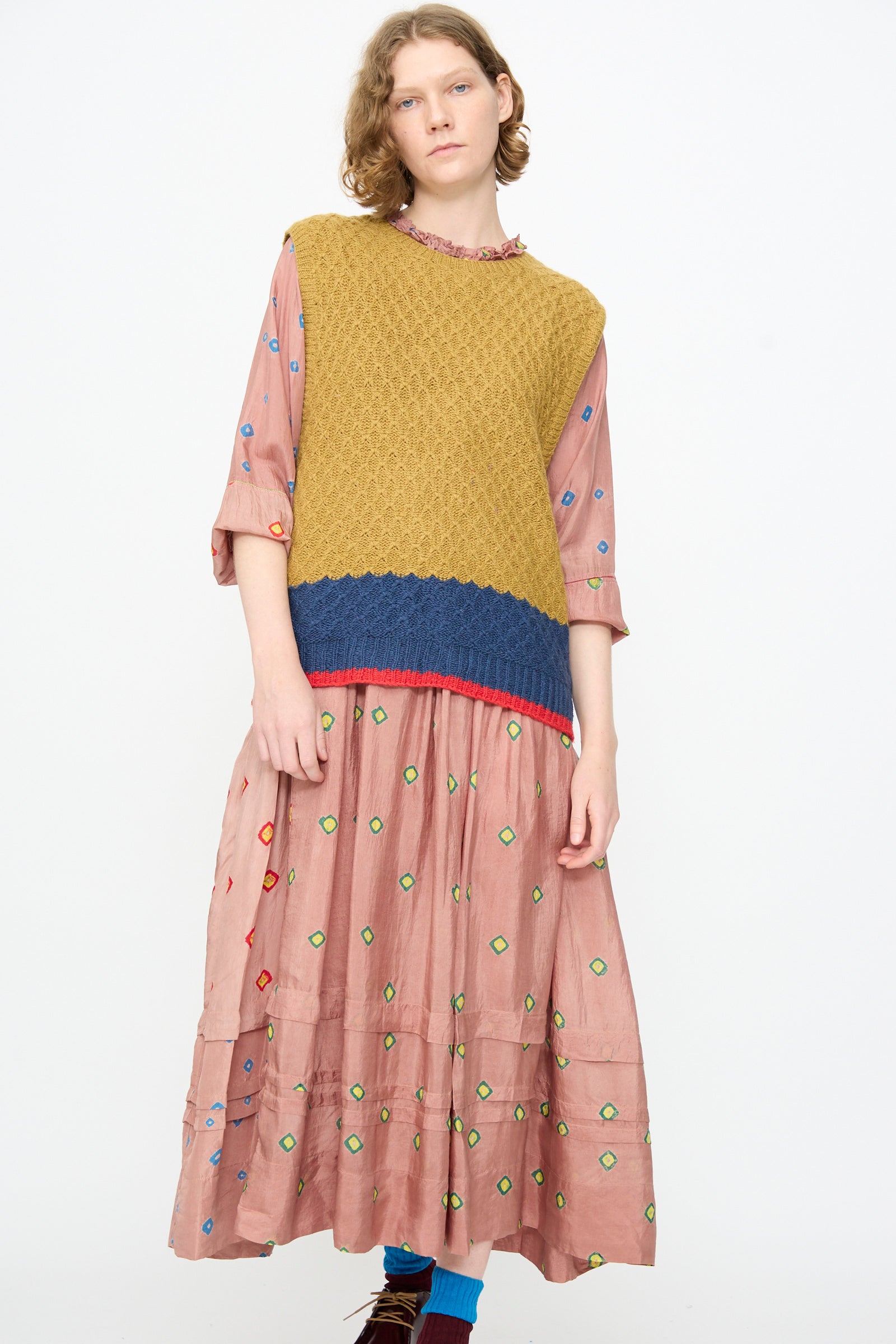 A person wearing Injiri’s Knitted Wool Sweater in Ochre and Blue—an ankle-length pink dress with colorful patterns layered beneath a boxy, textured, sleeveless knit sweater—stands against a plain white background.