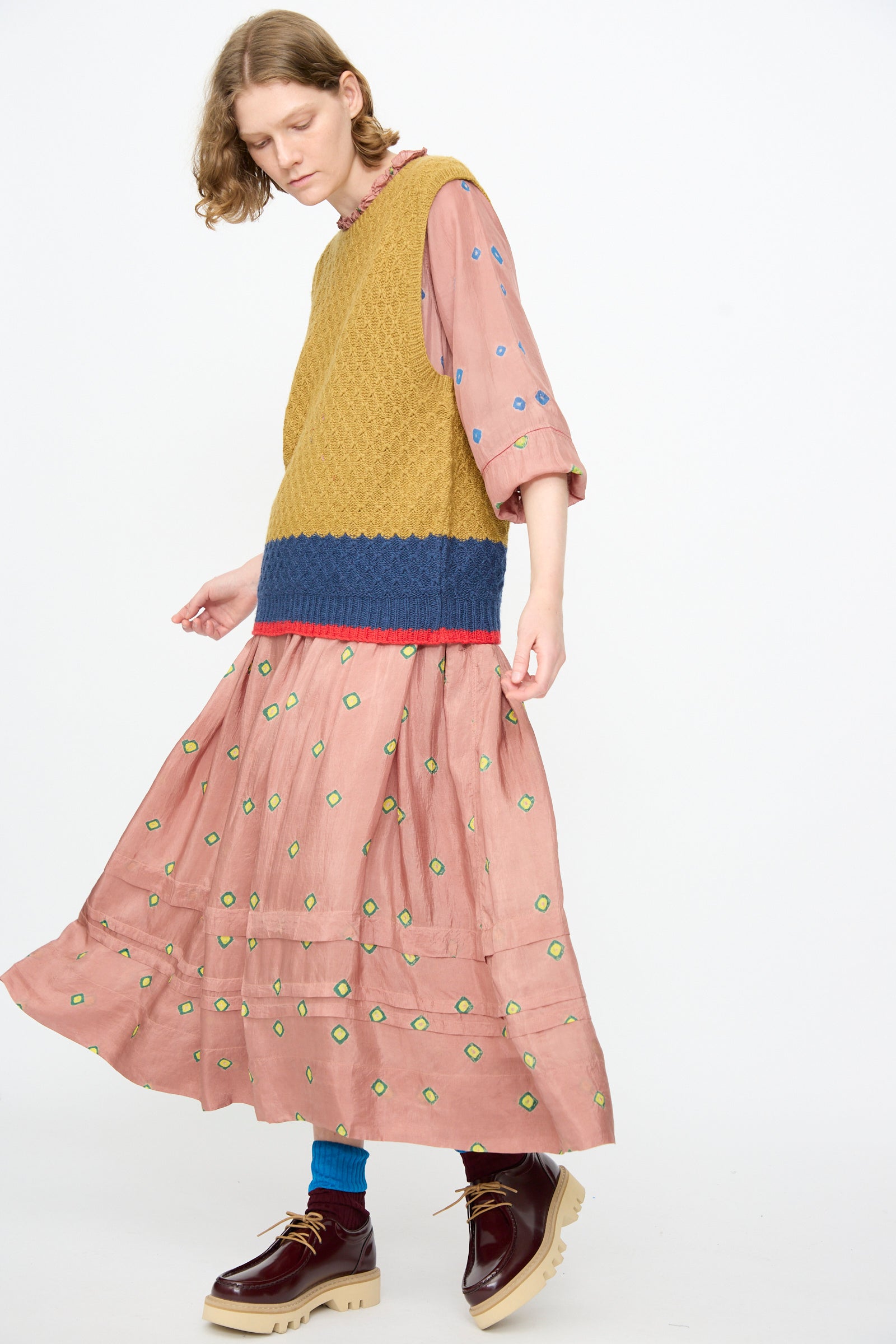 A person wearing a pink dress with green dots, Injiri’s Knitted Wool Sweater in Ochre and Blue with a boxy fit, blue socks, and burgundy chunky shoes stands against a plain white background.
