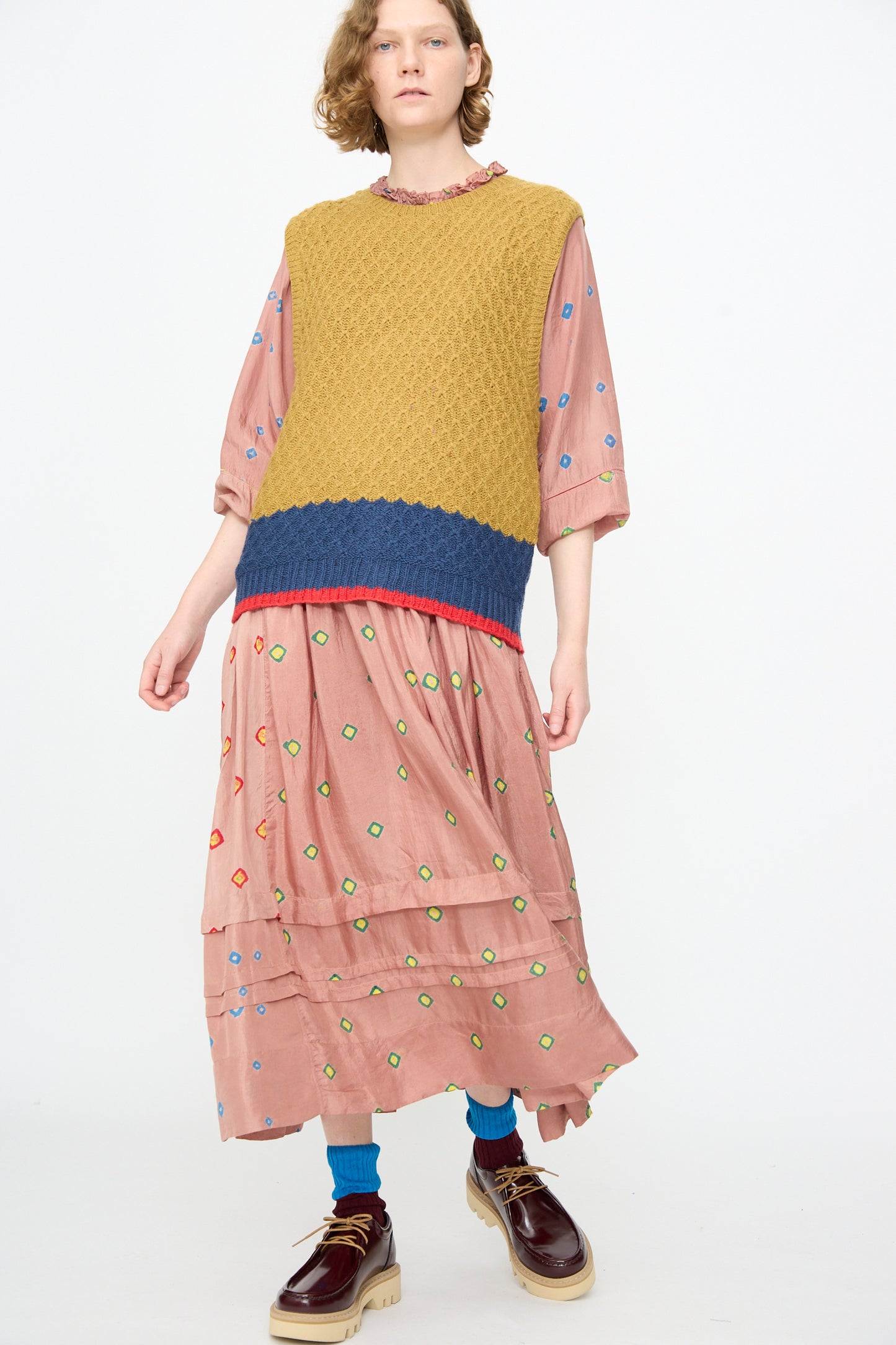 A person is wearing a pink dress with colorful dots, layered with the Injiri Knitted Wool Sweater in Ochre and Blue—a boxy fit sleeveless knit featuring blue trim—along with blue socks and brown platform shoes, standing against a plain white background.
