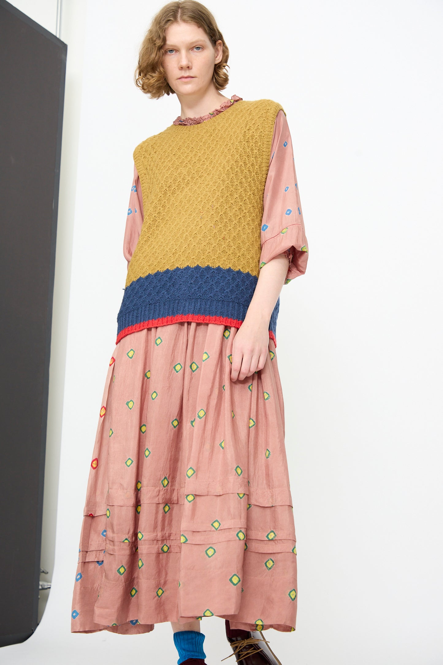 A person wears the Injiri Knitted Wool Sweater in Ochre and Blue, a textured, sleeveless knit with a boxy fit, layered over a loose pink dress adorned with multicolored geometric patterns. The look is completed with blue socks and brown shoes, set against a plain white background.