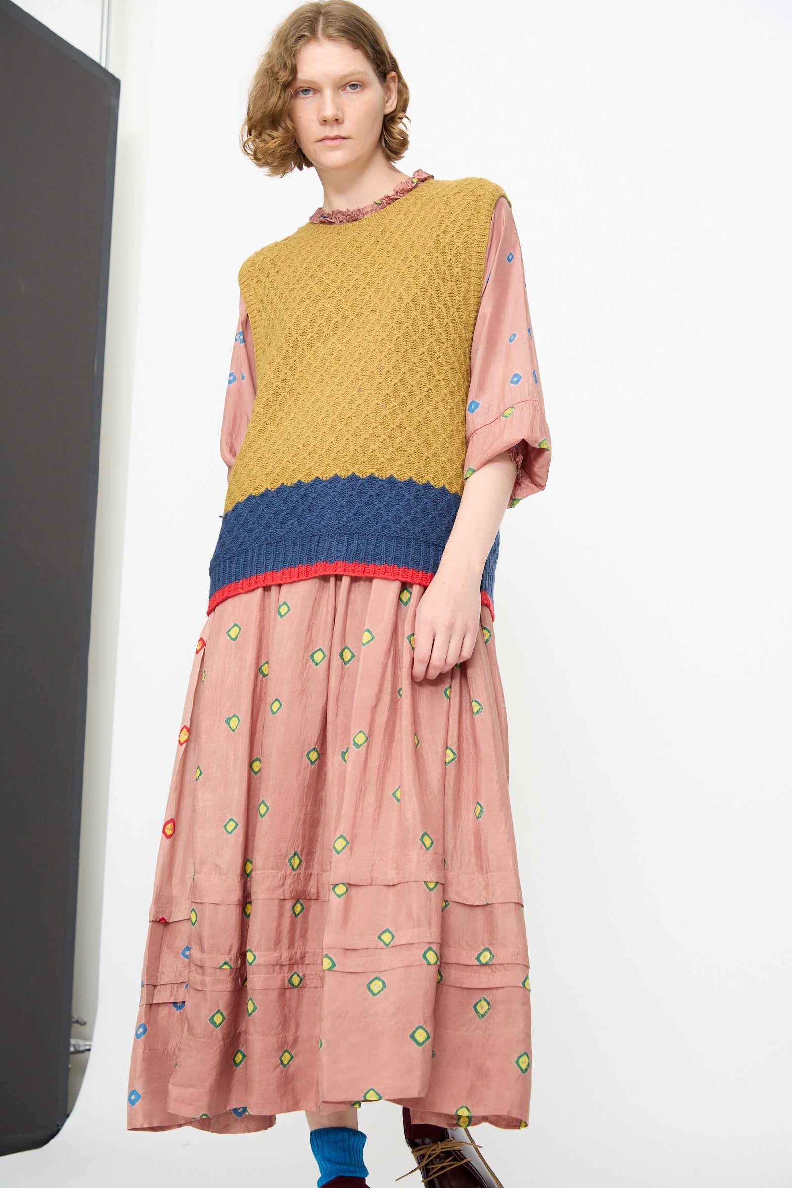 A person wears the Injiri Knitted Wool Sweater in Ochre and Blue, a textured, sleeveless knit with a boxy fit, layered over a loose pink dress adorned with multicolored geometric patterns. The look is completed with blue socks and brown shoes, set against a plain white background.