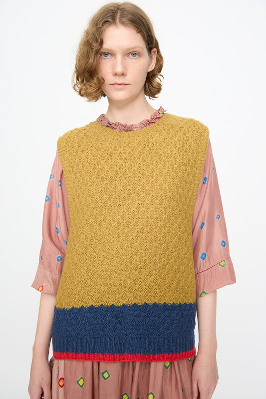 A person wears the Injiri Knitted Wool Sweater in Ochre and Blue, a boxy-fit, sleeveless knit sweater, layered over a pink dress with colorful embroidered shapes, standing against a plain white background.