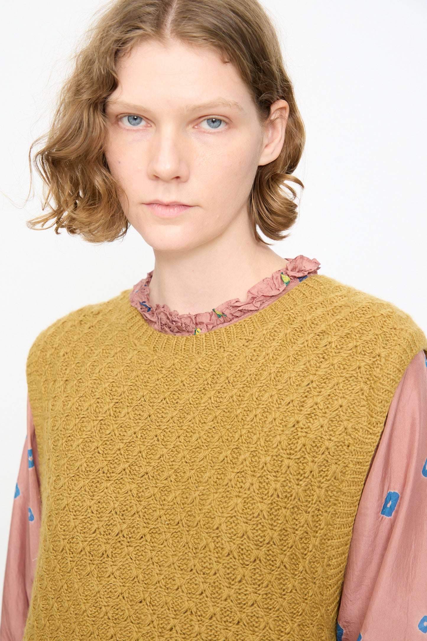 A person with short wavy hair is wearing the Injiri Knitted Wool Sweater in Ochre and Blue—a boxy fit, sleeveless knit vest—layered over a pink ruffled blouse with blue accents, standing against a plain light background.