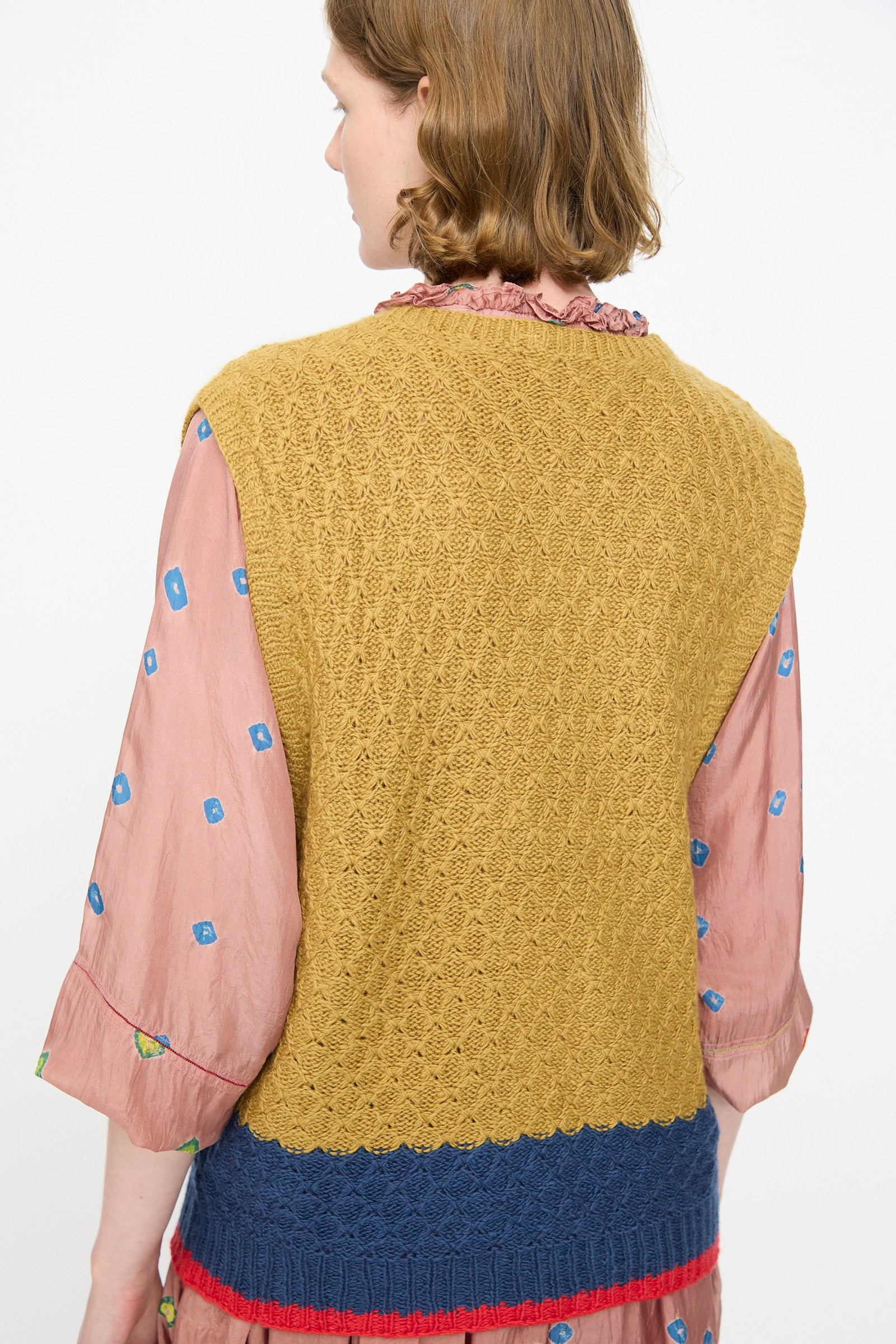 A person with wavy, light brown hair is seen from behind wearing the Injiri Knitted Wool Sweater in Ochre and Blue—a sleeveless, textured piece—layered over a pink blouse featuring blue square patterns.