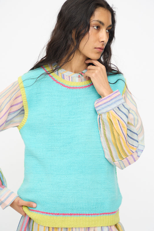 A person wearing the Injiri Kulluwool Sweater Vest in Aqua over a pastel striped blouse stands against a plain white background, gazing to the side with one hand near their chin.