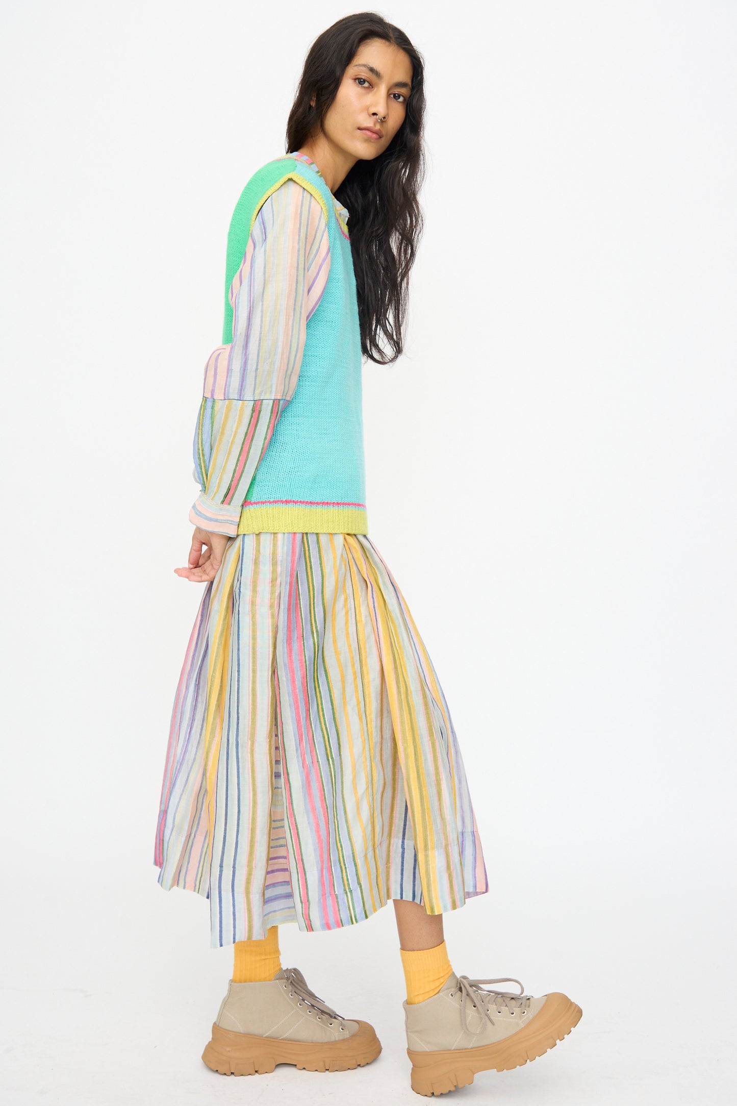 A woman in profile wears an Injiri Kulluwool Sweater Vest in Aqua over a pastel striped dress, paired with yellow socks and beige platform shoes, set against a plain white background.