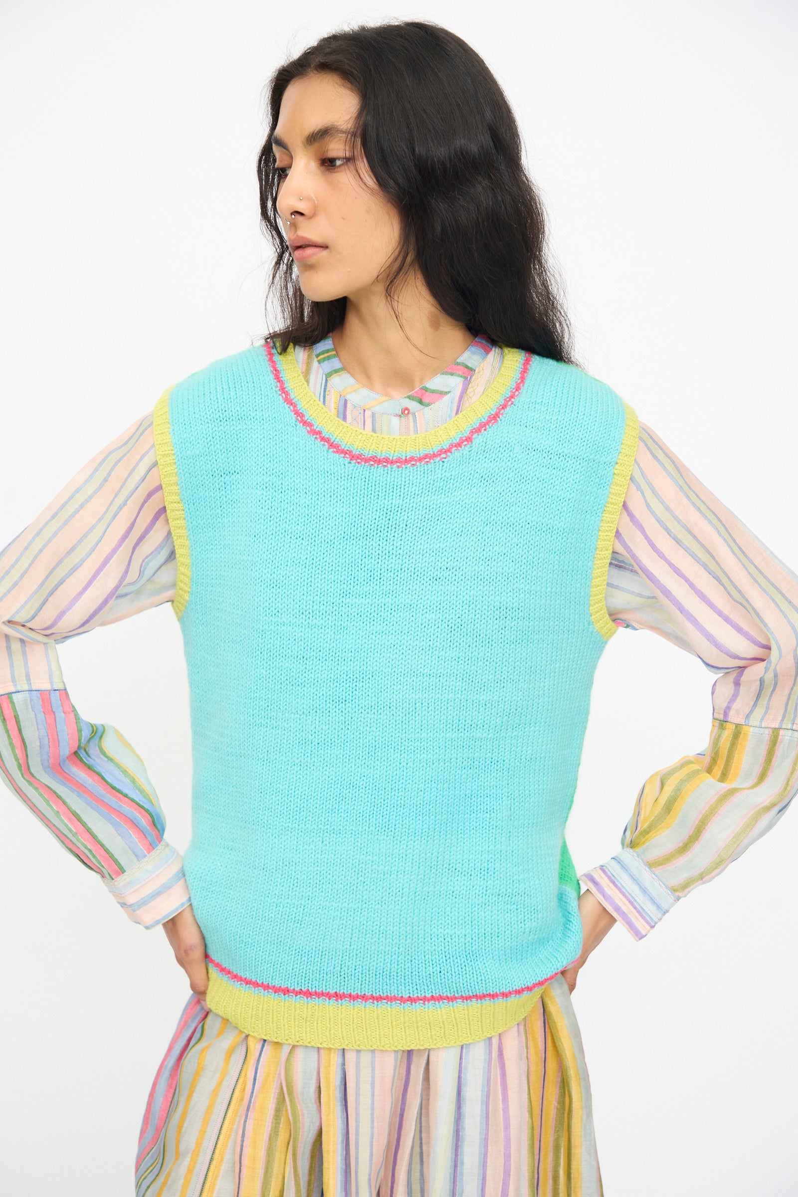 A person wears the Injiri Kulluwool Sweater Vest in Aqua, a sleeveless 100% wool design, over a pastel striped long-sleeve dress and stands with hands on hips, looking to the side.