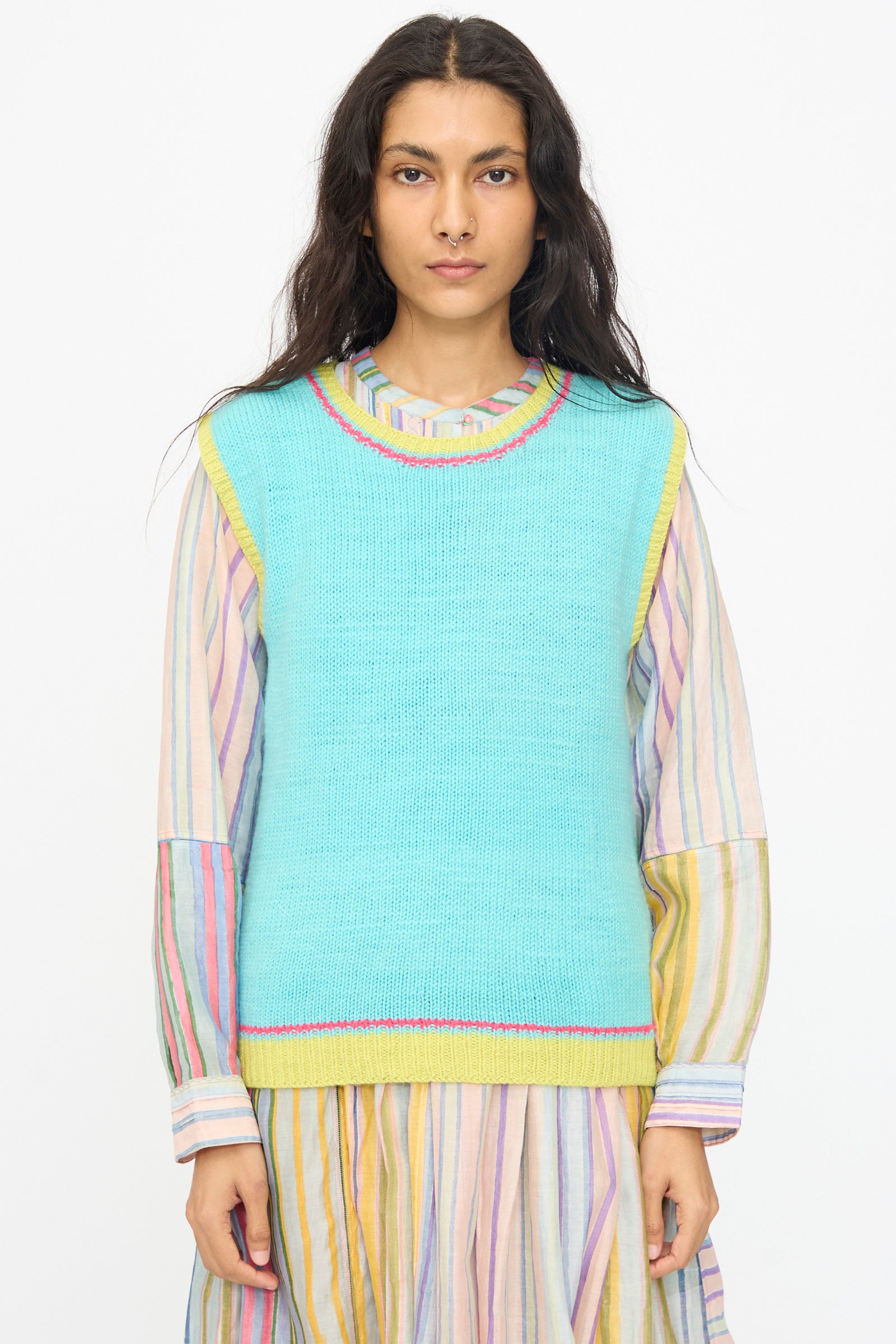 A person wears the Injiri Kulluwool Sweater Vest in Aqua over a pastel-striped long-sleeve dress, standing facing forward against a plain background.