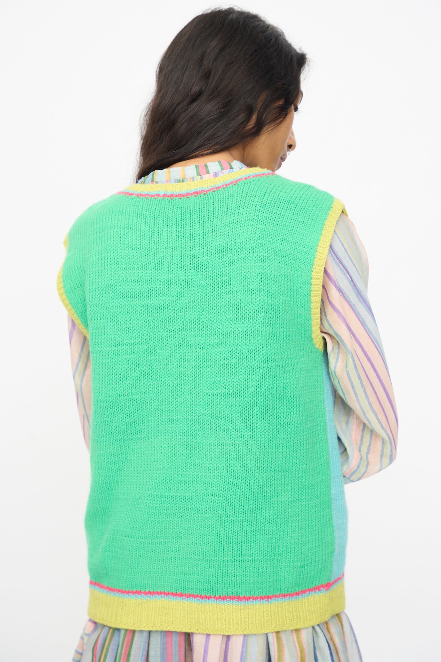 A person with long dark hair wears the Injiri Kulluwool Sweater Vest in Aqua, styled over a pastel striped shirt and photographed from the back.