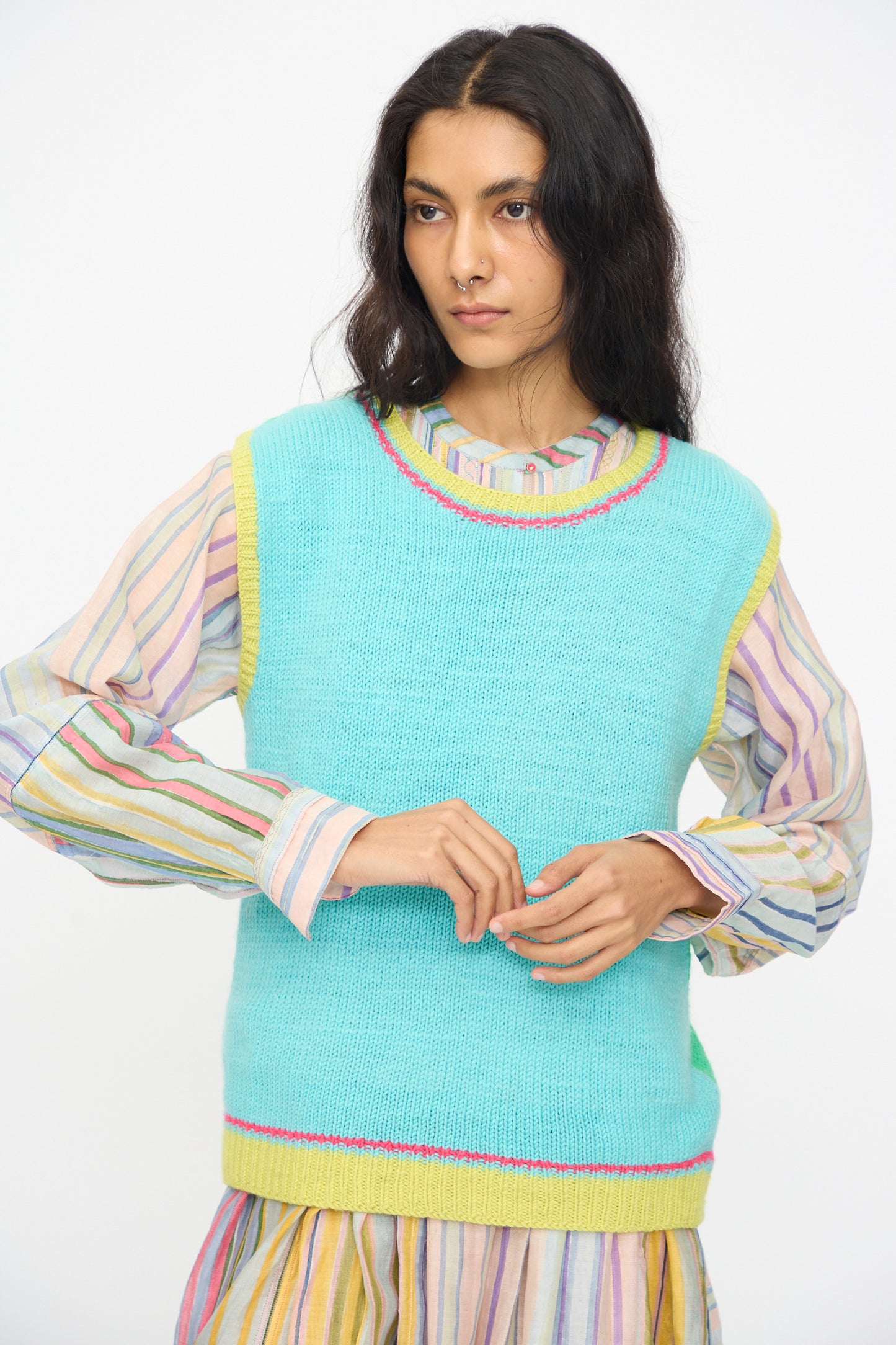 A woman in a pastel striped dress and the Injiri Kulluwool Sweater Vest in Aqua, crafted from 100% wool and detailed with yellow and pink trim, stands against a plain white background.