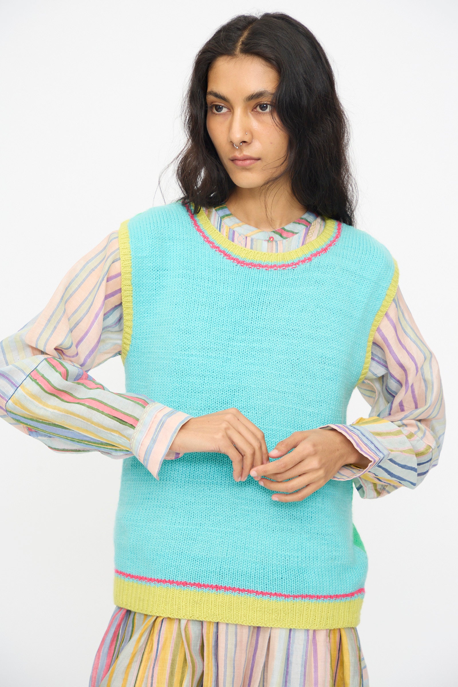 A woman in a pastel striped dress and the Injiri Kulluwool Sweater Vest in Aqua, crafted from 100% wool and detailed with yellow and pink trim, stands against a plain white background.