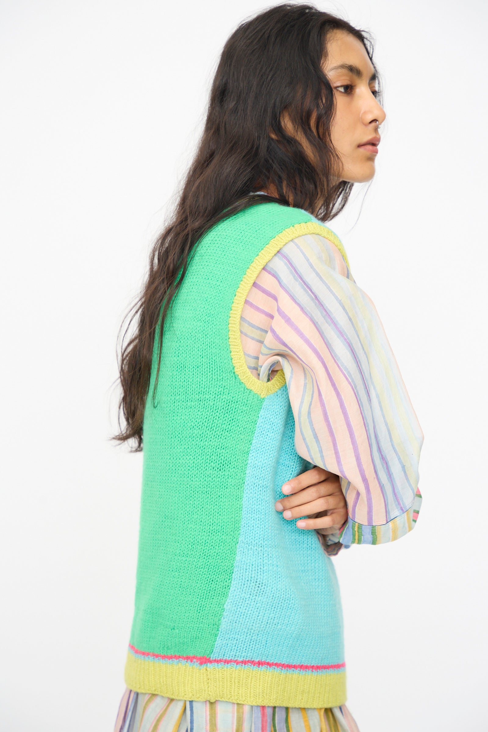 Person with long dark hair wears the Injiri Kulluwool Sweater Vest in Aqua over a pastel striped blouse, standing against a plain white background.