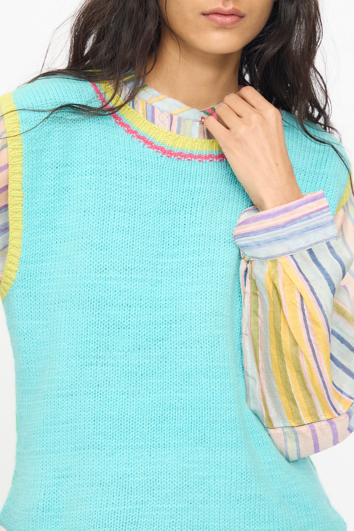 A person wears the Injiri Kulluwool Sweater Vest in Aqua over a long-sleeve, multi-colored striped shirt, with only their lower face and upper body visible.