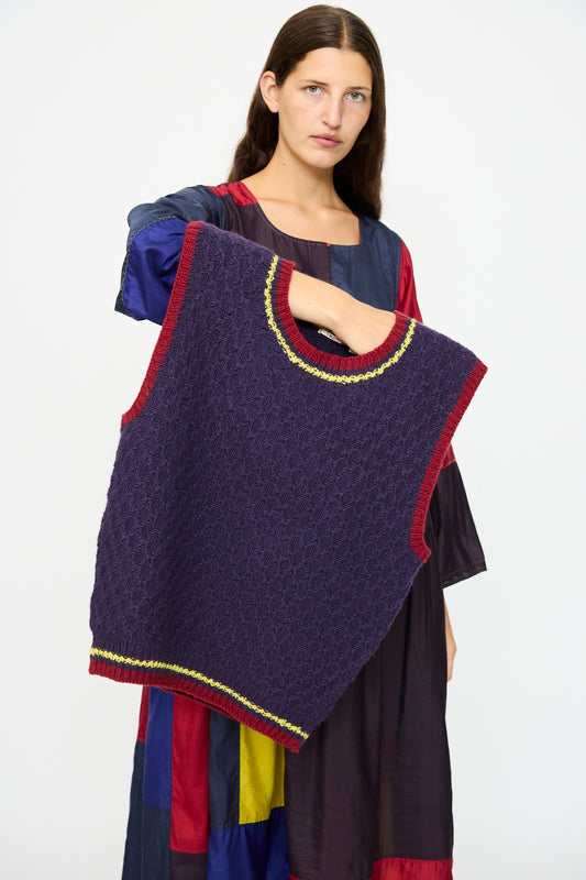 A person holds up the Injiri Merino Wool Sweater Vest in Purple, Red and Yellow, while wearing a long-sleeved dress with a bold red, blue, yellow, and black color block pattern.