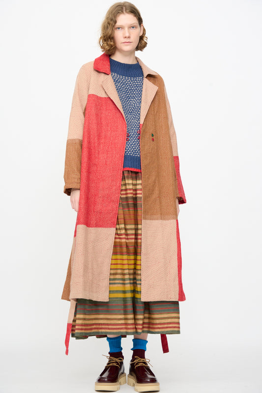 A person wearing the Injiri Qasab Wool and Silk Jacket in Red and Brown Multi—a long patchwork coat crafted from a luxurious wool-silk blend—paired with a blue knit sweater, striped skirt, blue socks, and brown shoes, stands against a plain white background. This distinctive look is made with carefully sourced materials from India.